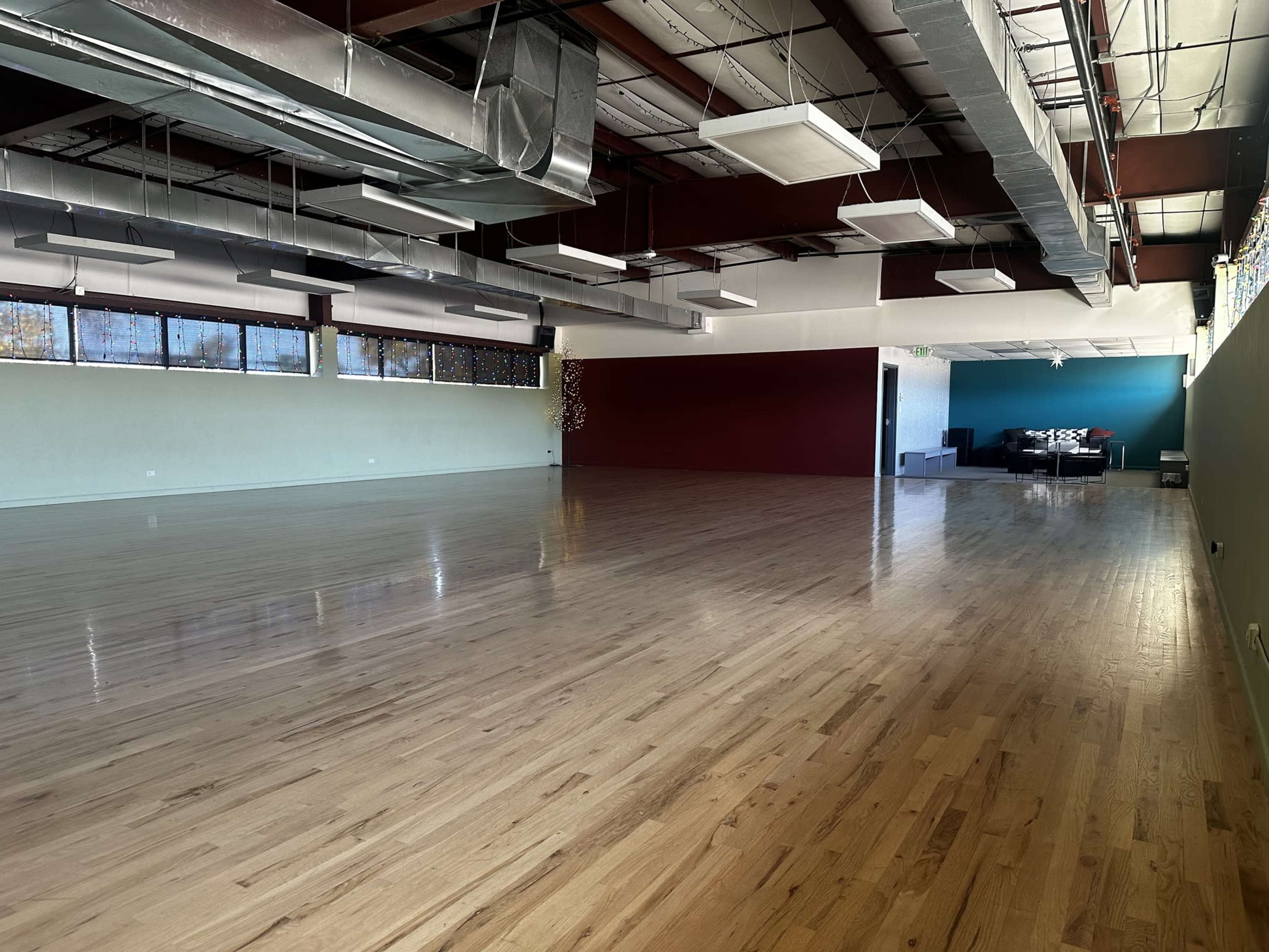 A spacious, empty studio with polished wooden floors and large windows, featuring a seating area in the back.
