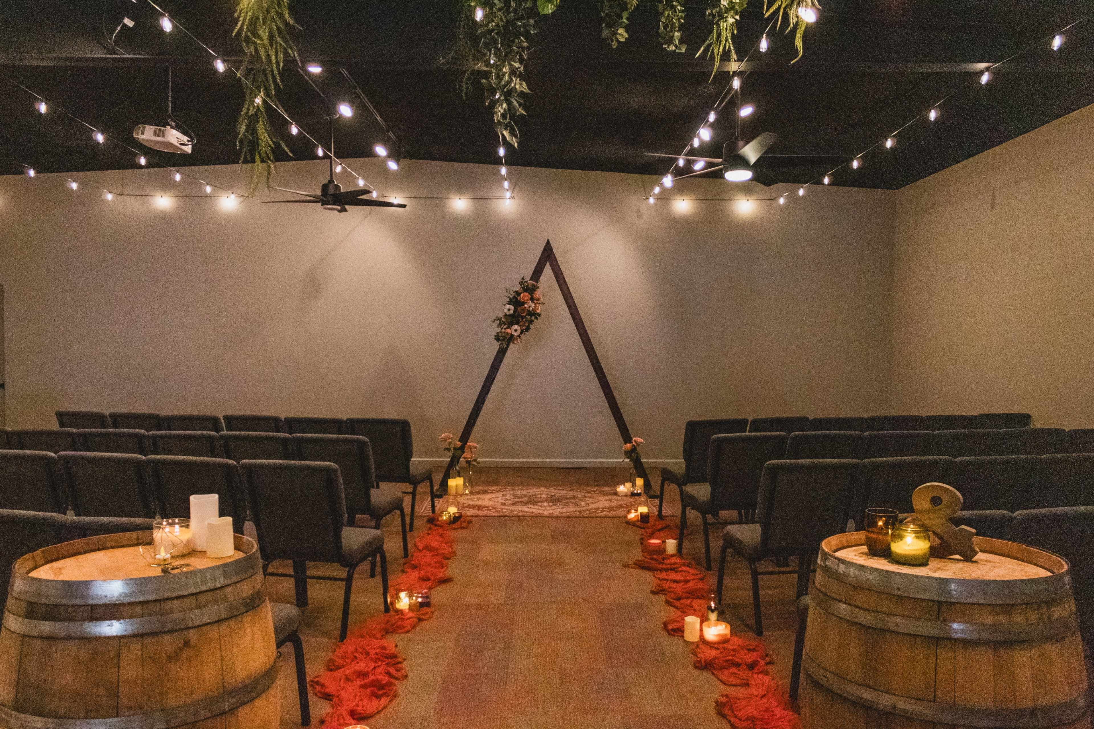 The image shows a wedding ceremony space with rows of chairs facing an altar adorned with flowers, surrounded by lit candles and scattered fabric, set against a backdrop with string lights.
