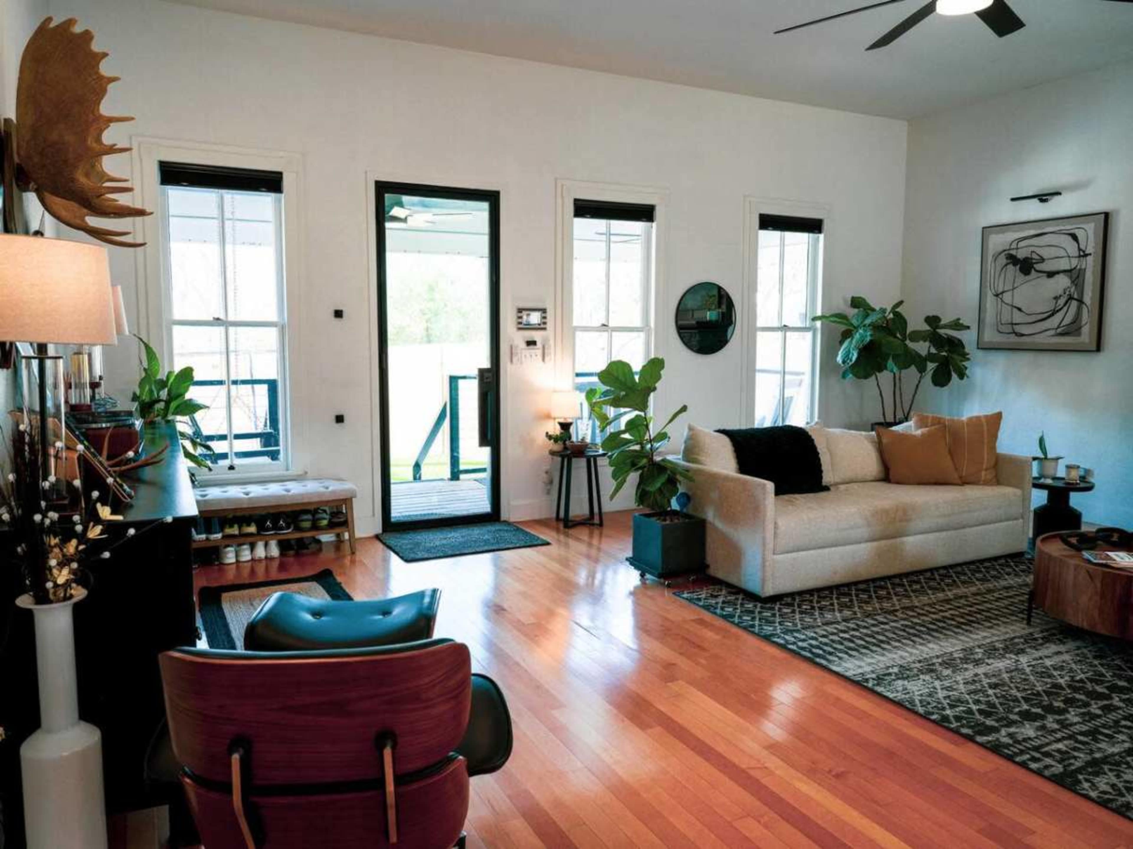 The image shows a bright living room with large windows, hardwood floors, a beige sofa, and various indoor plants.
