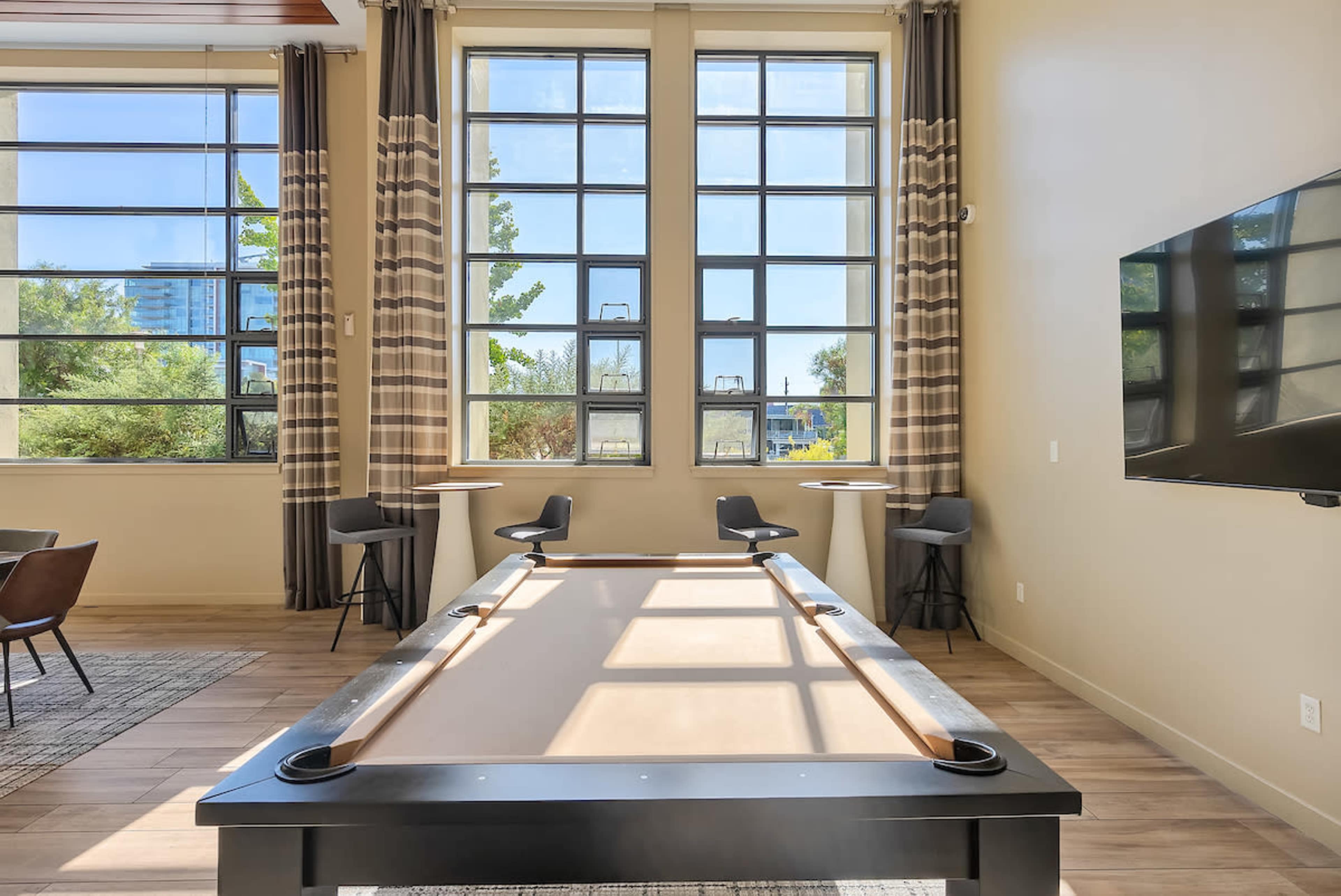 A pool table is positioned in a room with large windows, surrounded by several chairs and small tables.