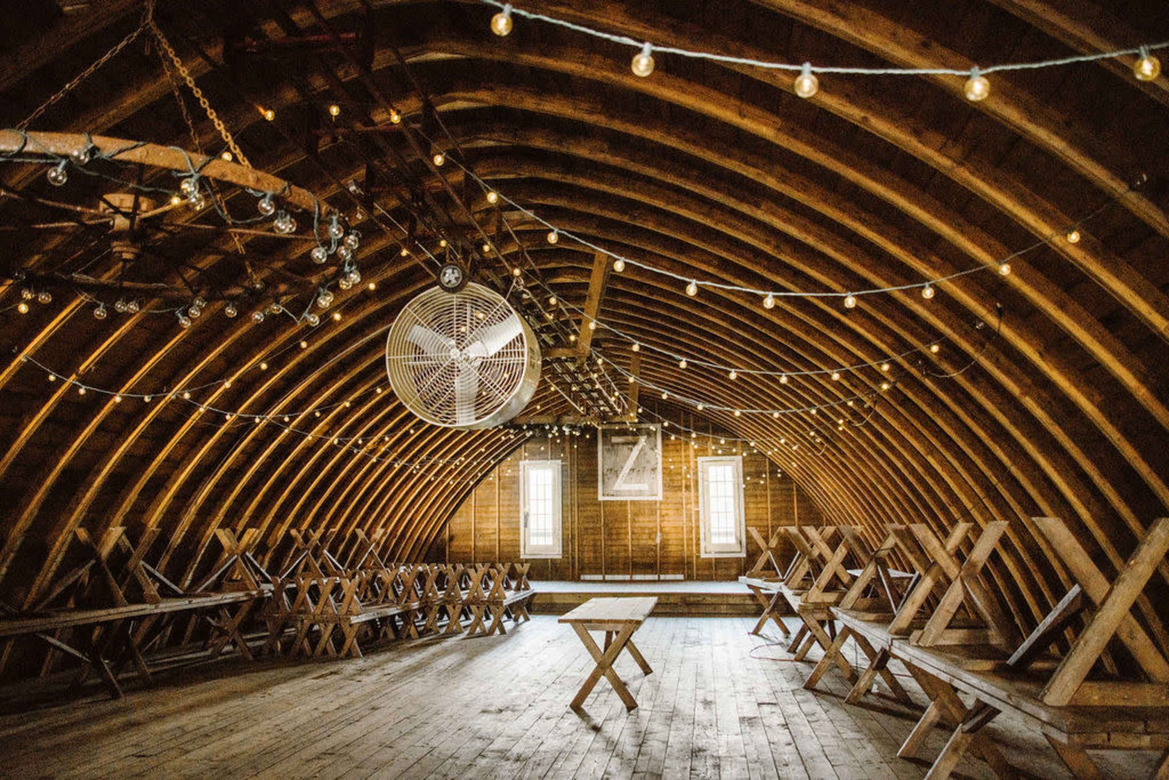 The interior of a rustic wooden structure features a high curved ceiling, string lights overhead, and several tables arranged throughout the space.