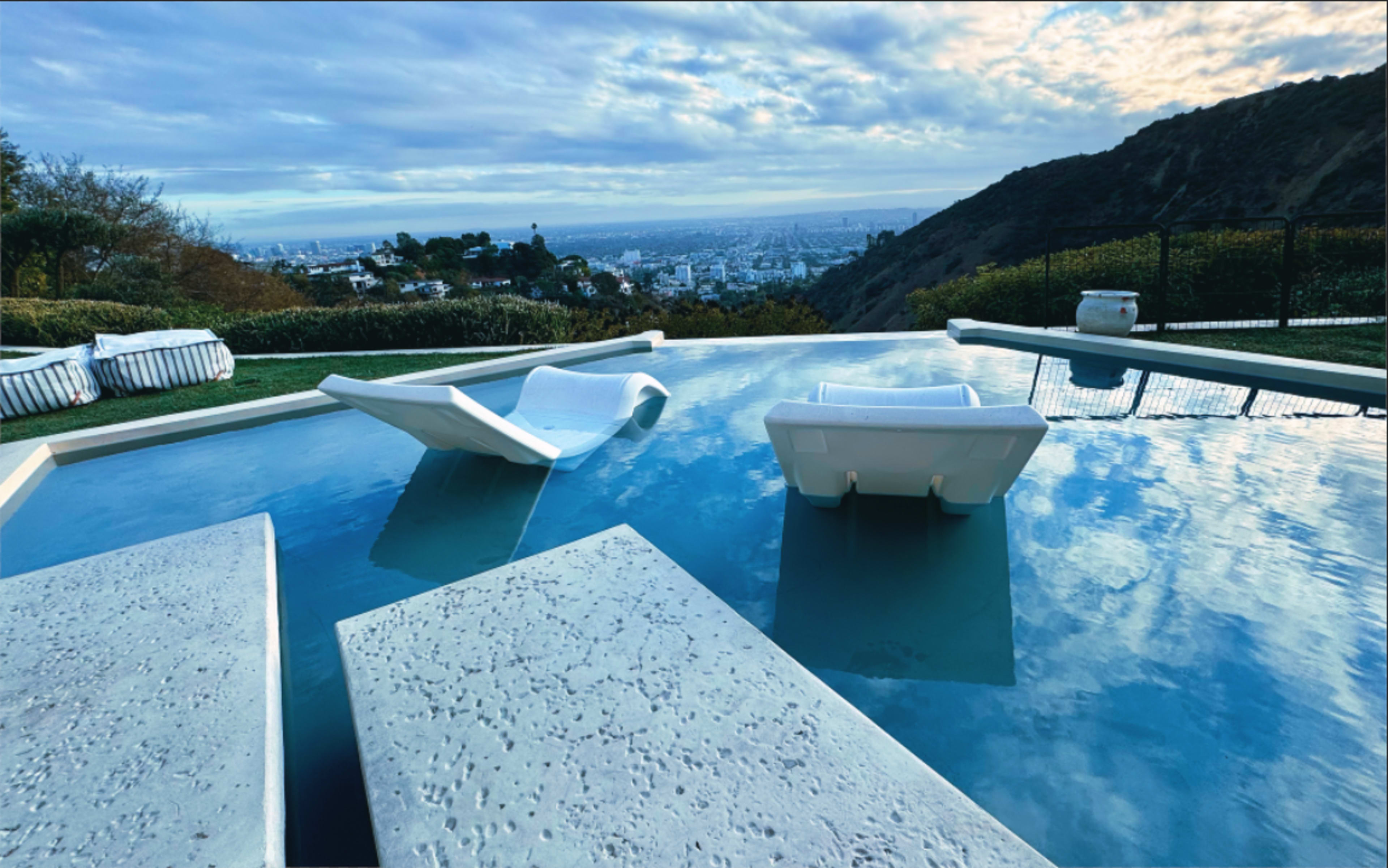 A view of a swimming pool with two lounge chairs partially submerged, overlooking a cityscape and mountains under a cloudy sky.