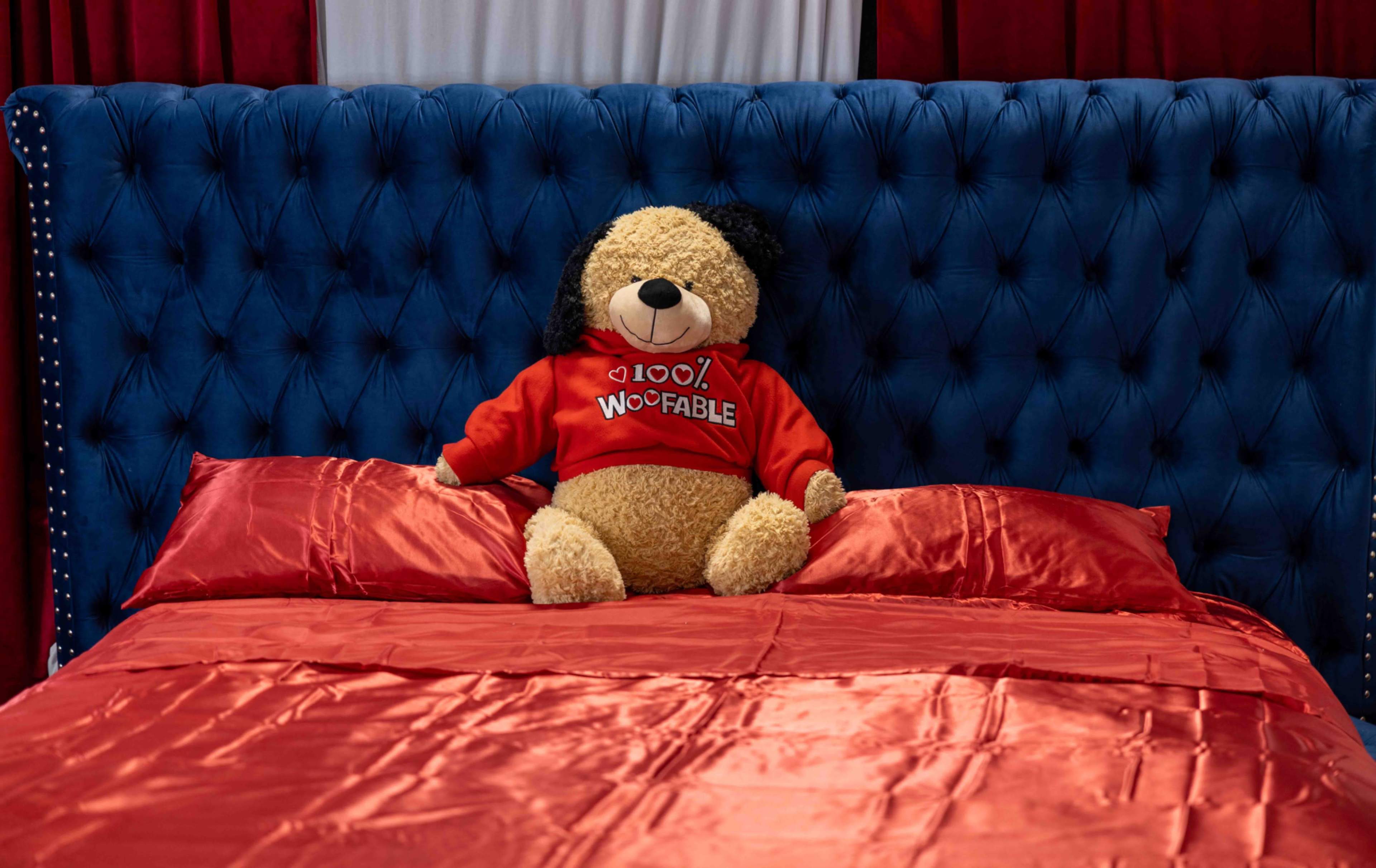 A large stuffed dog wearing a red sweater sits on a neatly made bed with shiny red bedding in a room with a blue tufted headboard.