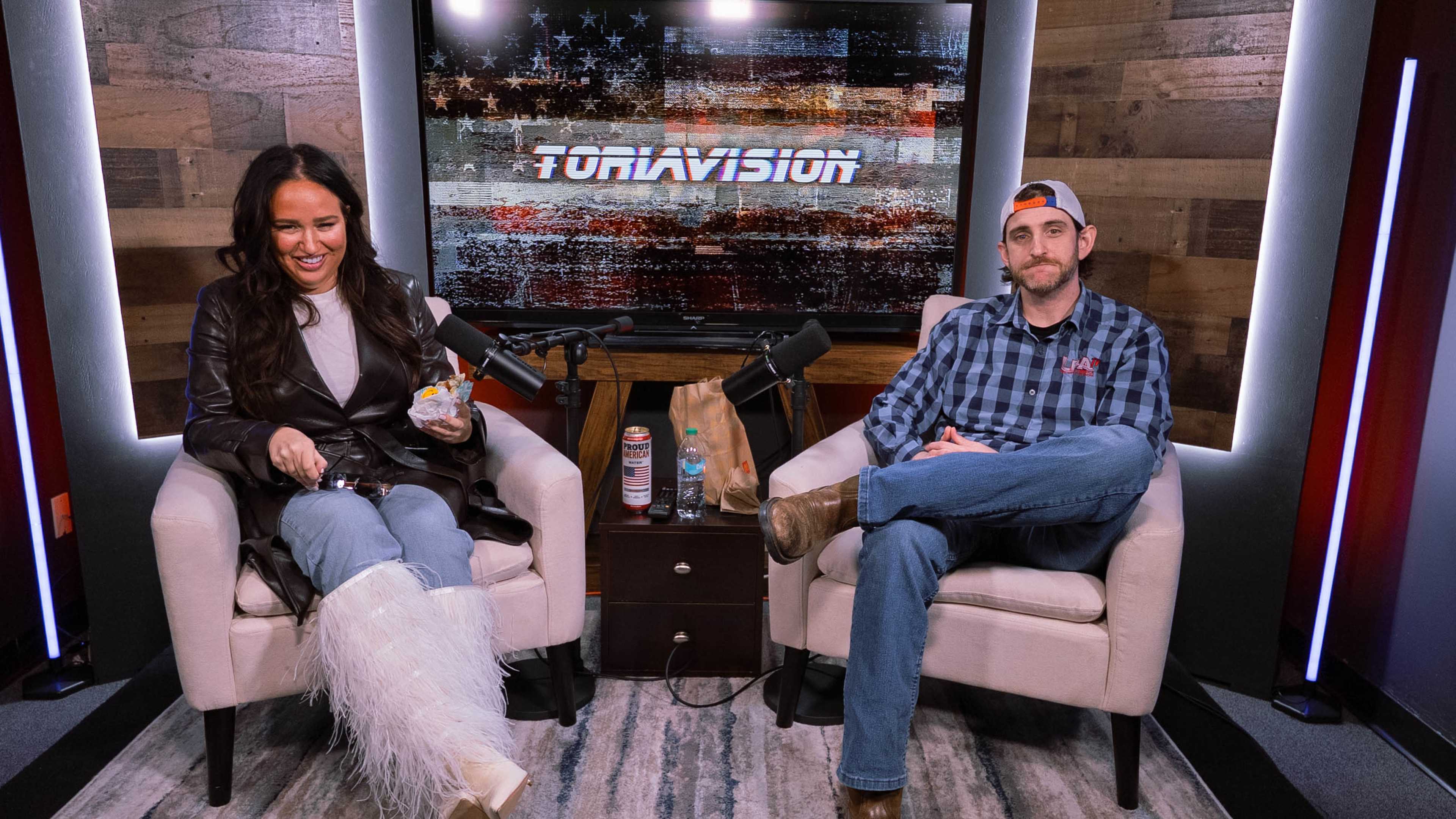 A man and a woman sit on chairs in a studio setting, with microphones in front of them and a backdrop displaying the word "TORAVISION."