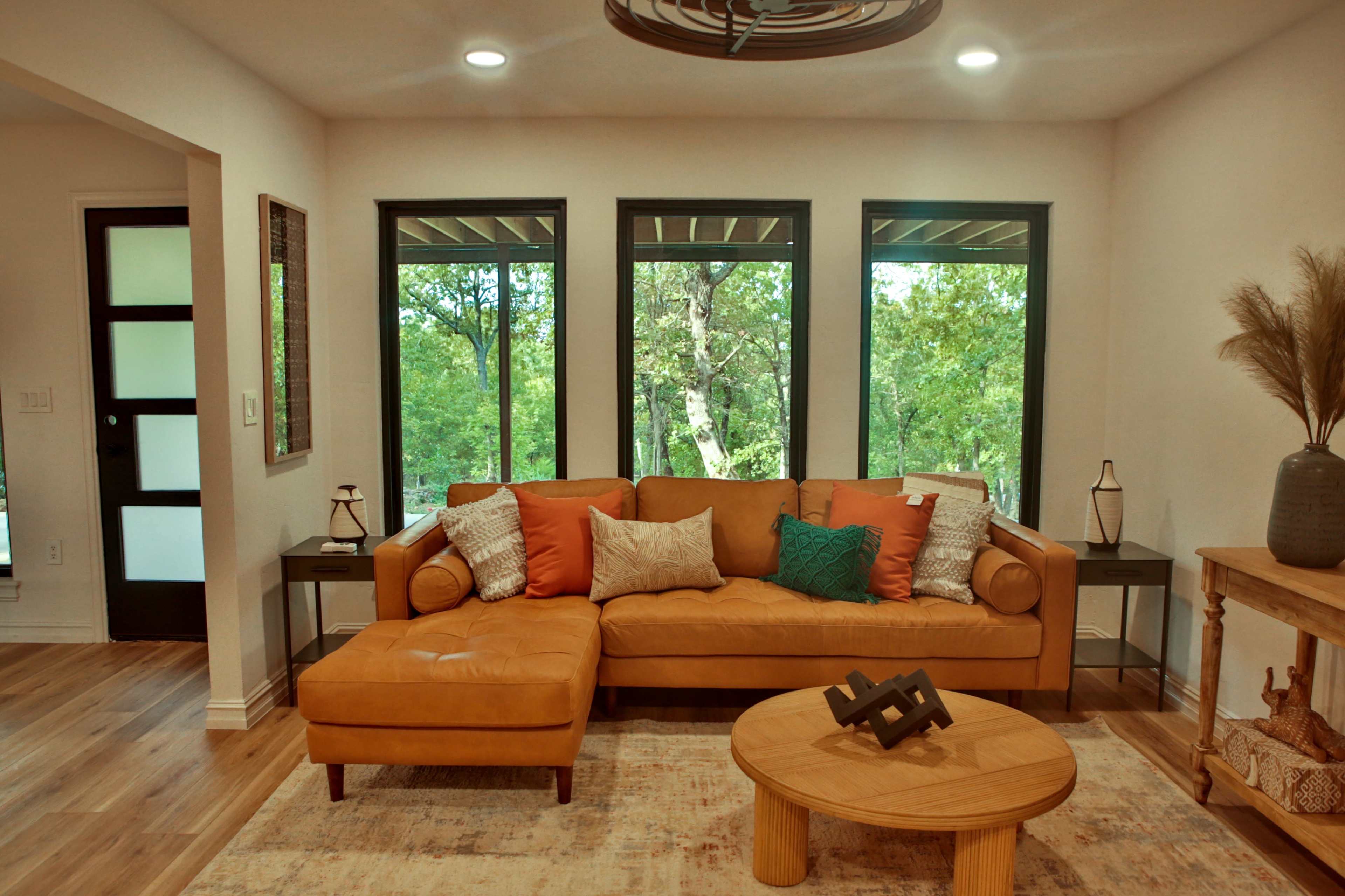A modern living room features a brown leather sofa with colorful throw pillows, two side tables, and large windows overlooking greenery.