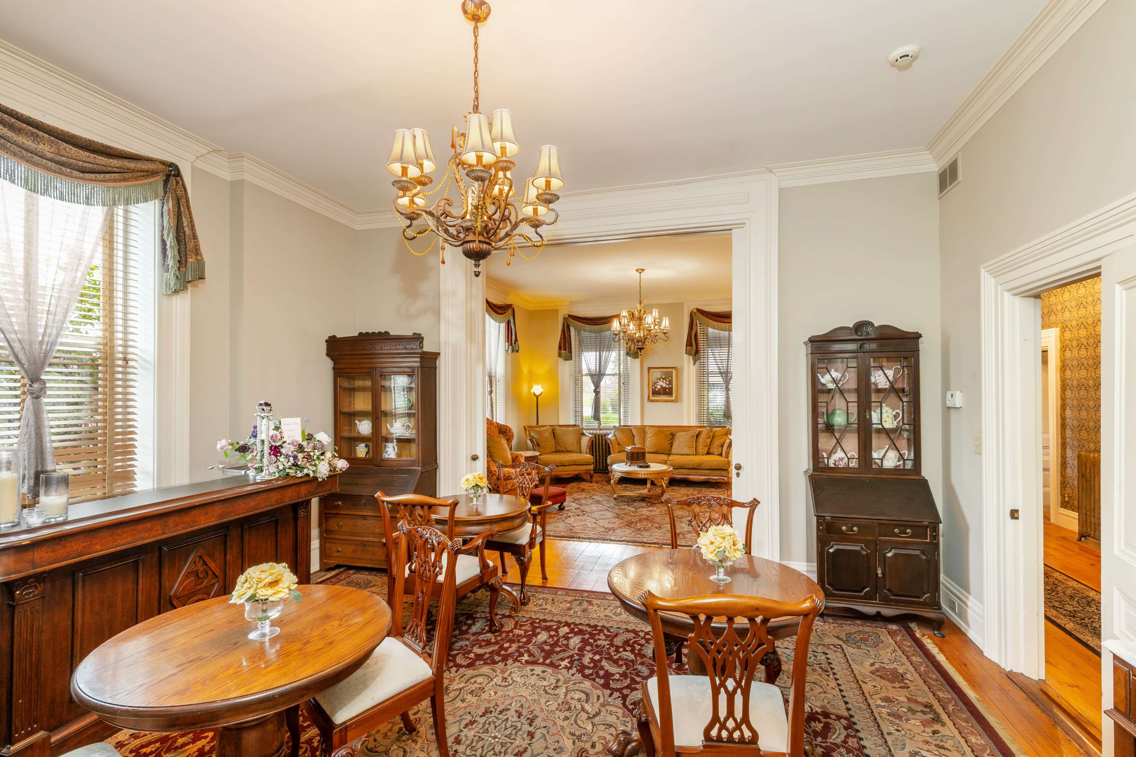 A well-furnished room features ornate light fixtures, wooden tables, and a view into a living area with a plush seating arrangement.