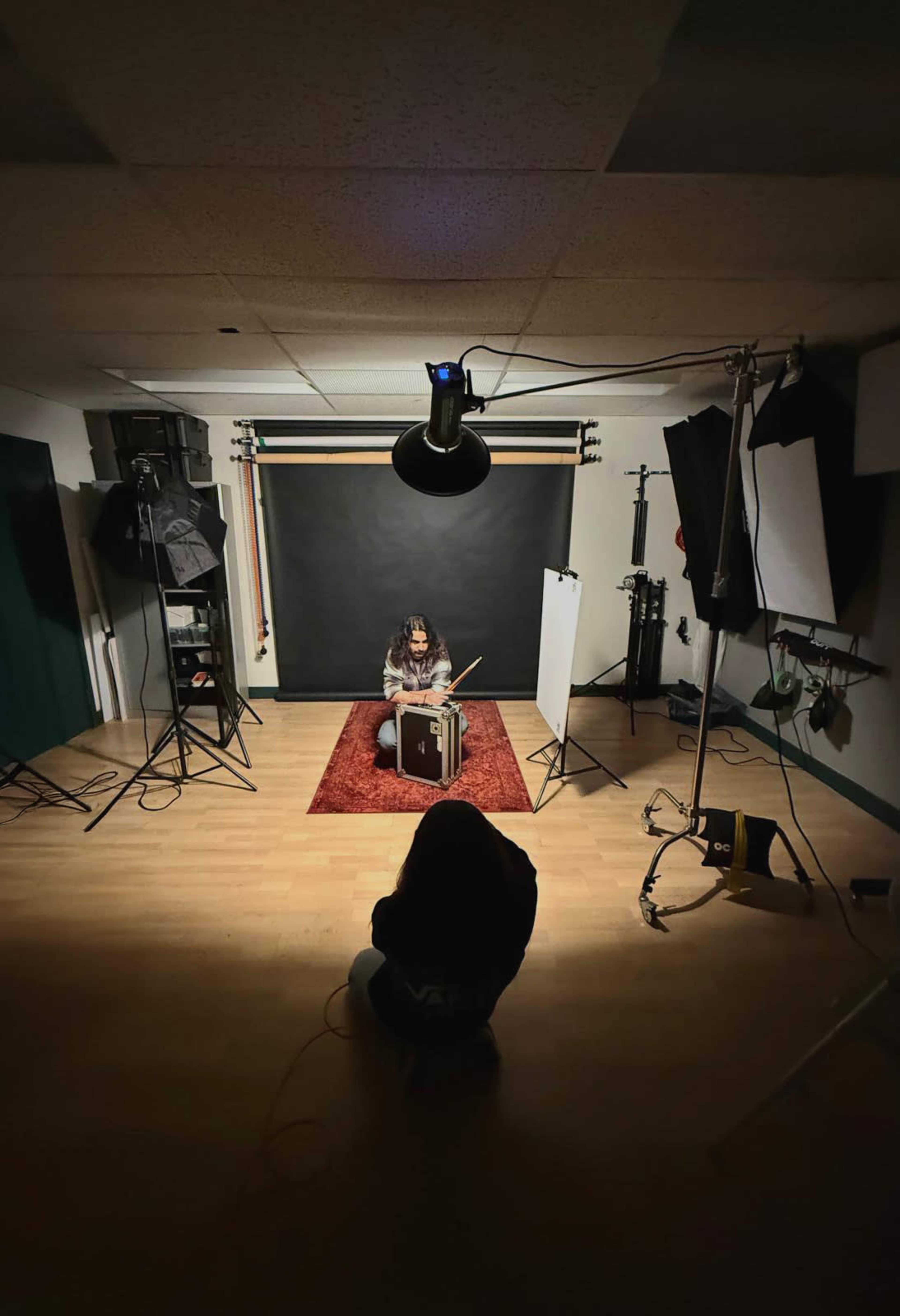 A person sits on a rug playing drums in a photography studio with various lighting equipment set up around them.