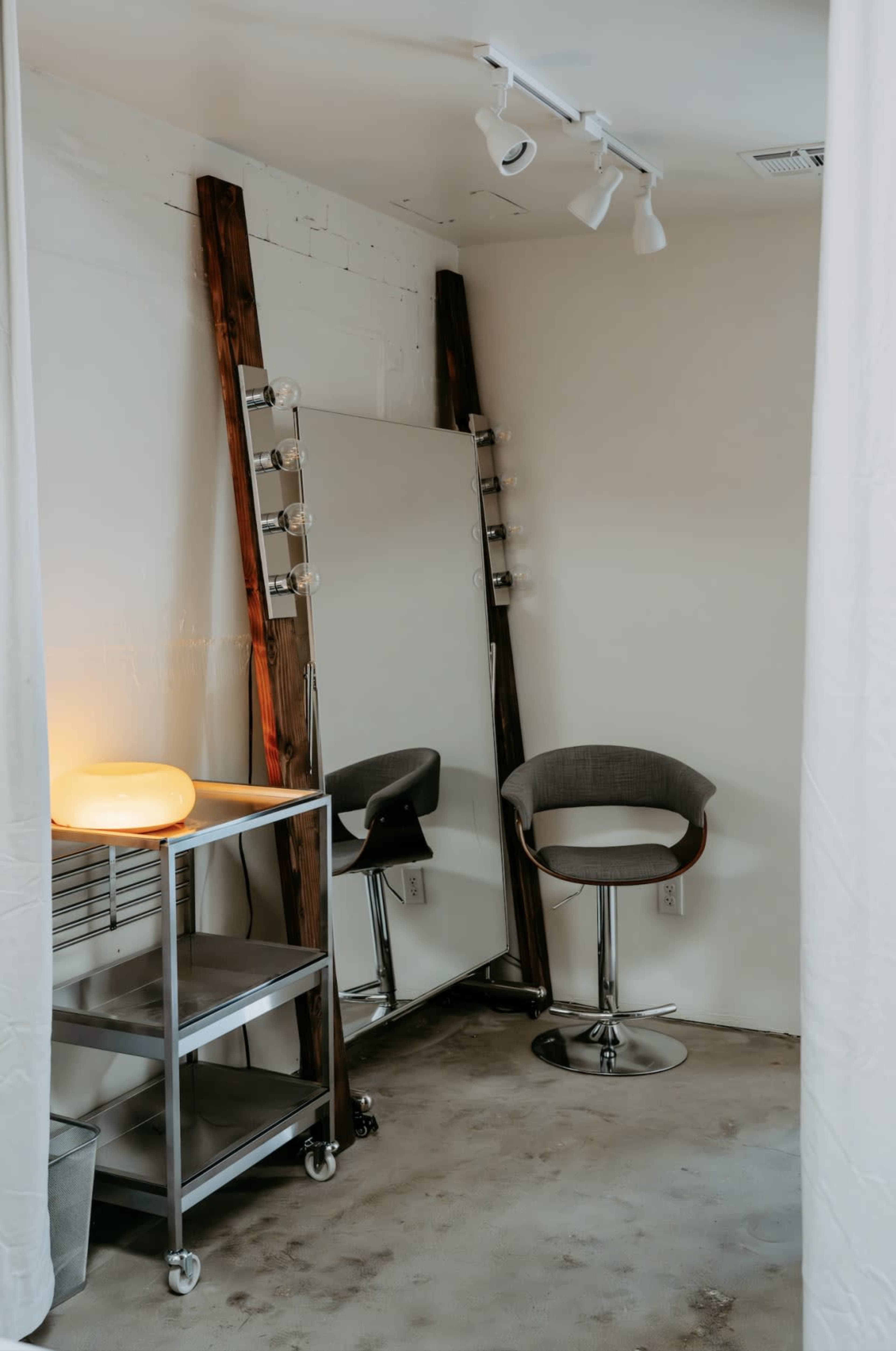 A well-lit corner of a room features a full-length mirror flanked by light bulbs, with two barstools and a mobile cart nearby.