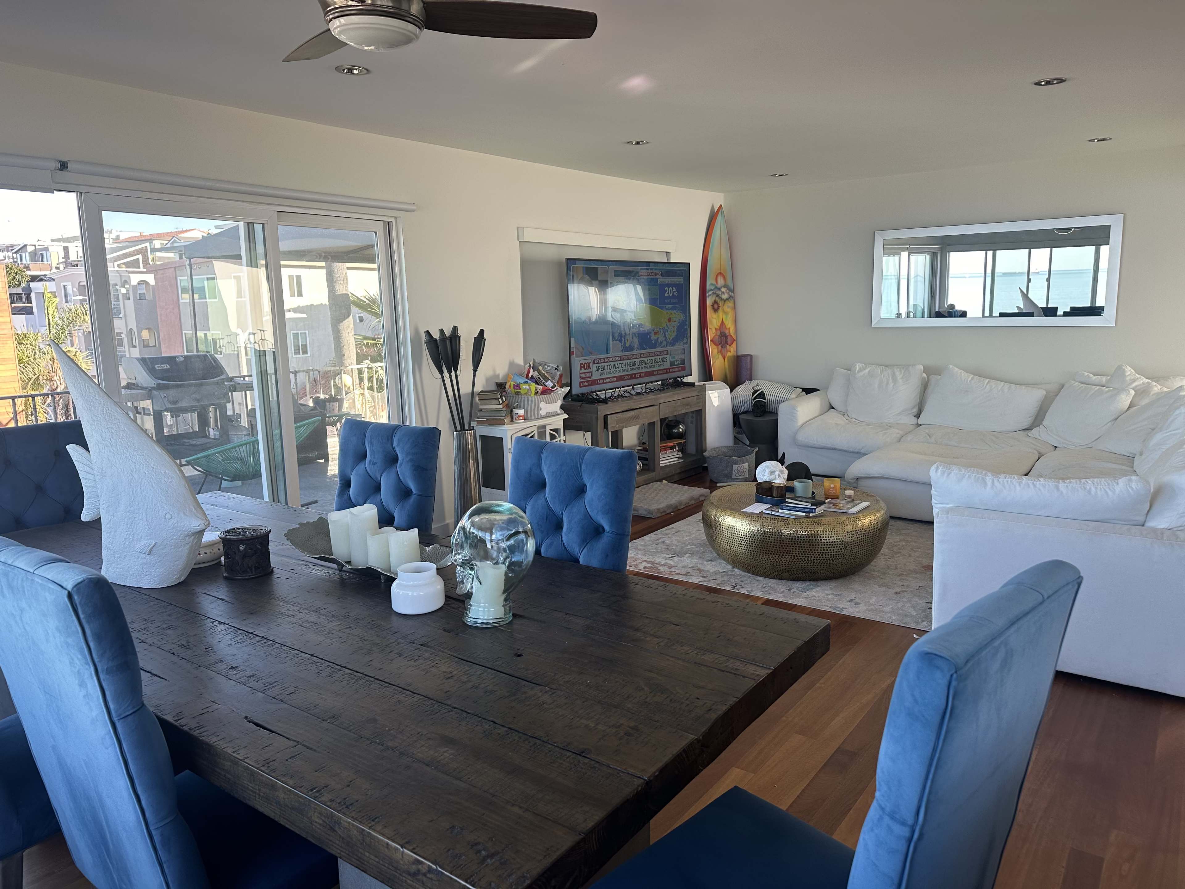 A bright living space features a wooden dining table with blue chairs, a white sectional sofa, a television on a stand, and large windows with a view outside.