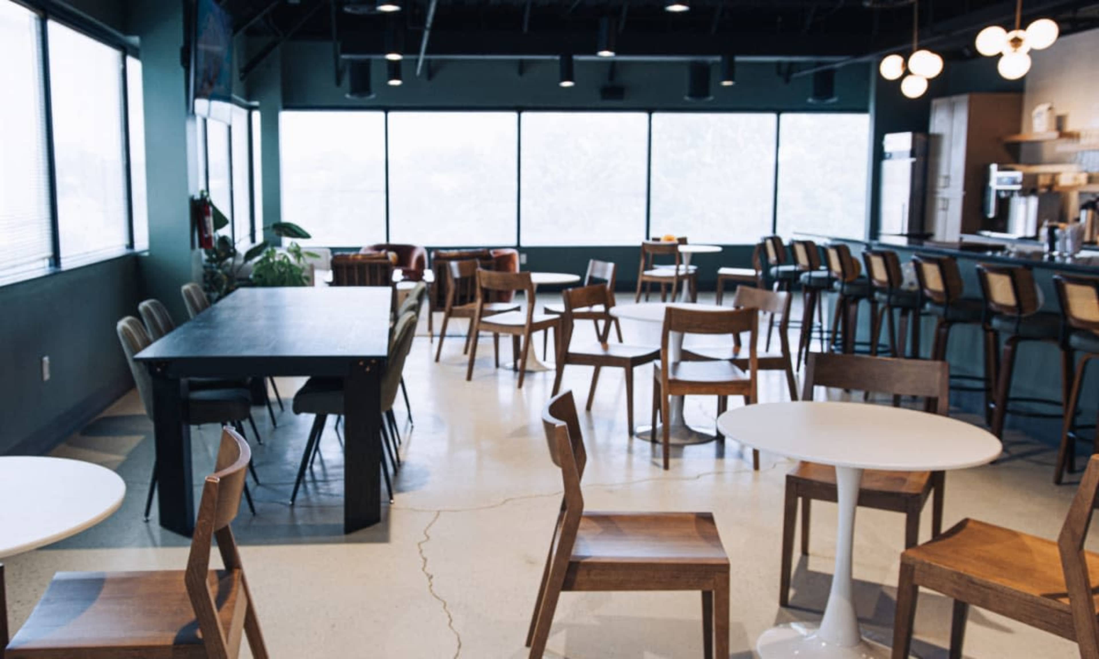 The image shows a spacious café or break room with various tables and chairs, large windows letting in natural light, and a kitchen area in the background.