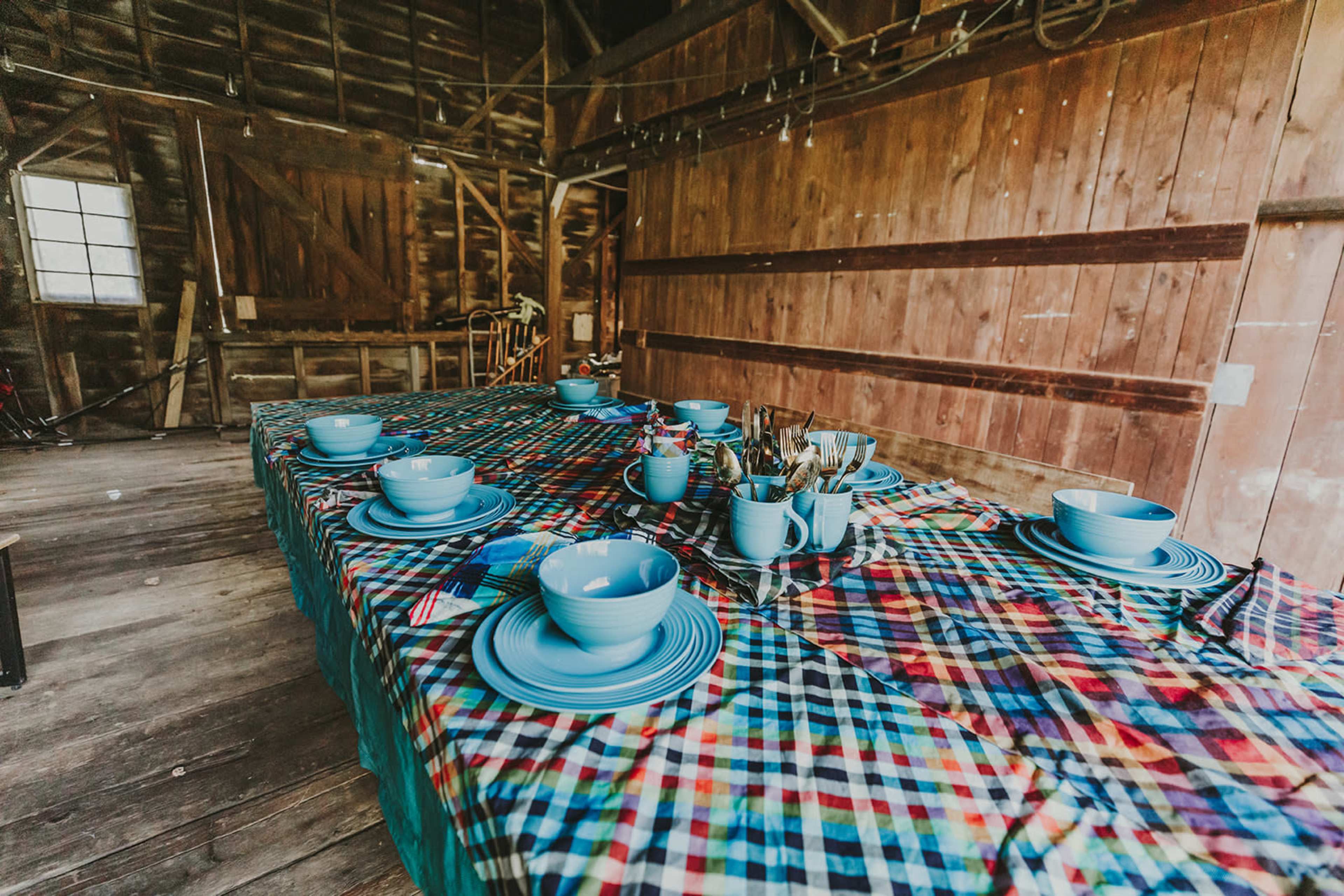 A long table covered with a plaid tablecloth is set with blue dishes and utensils in a rustic wooden interior.