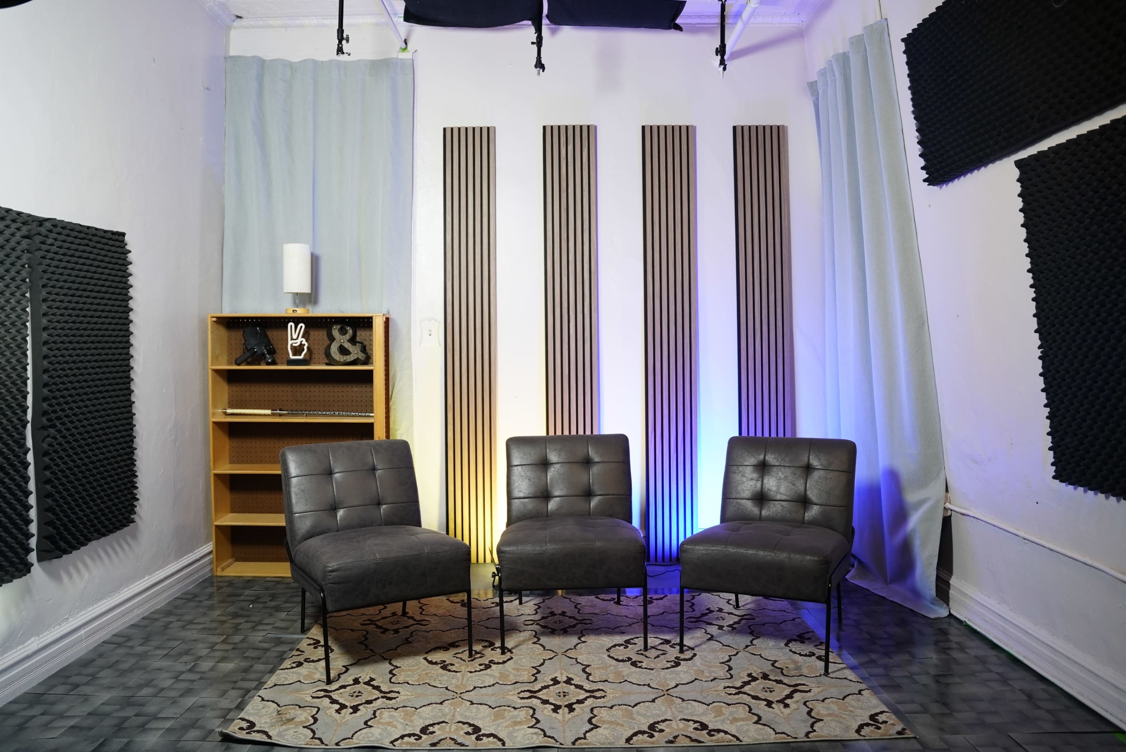 The image shows a small room with three gray chairs arranged in front of a bookshelf and acoustic panels on the walls, illuminated by soft colored lights.