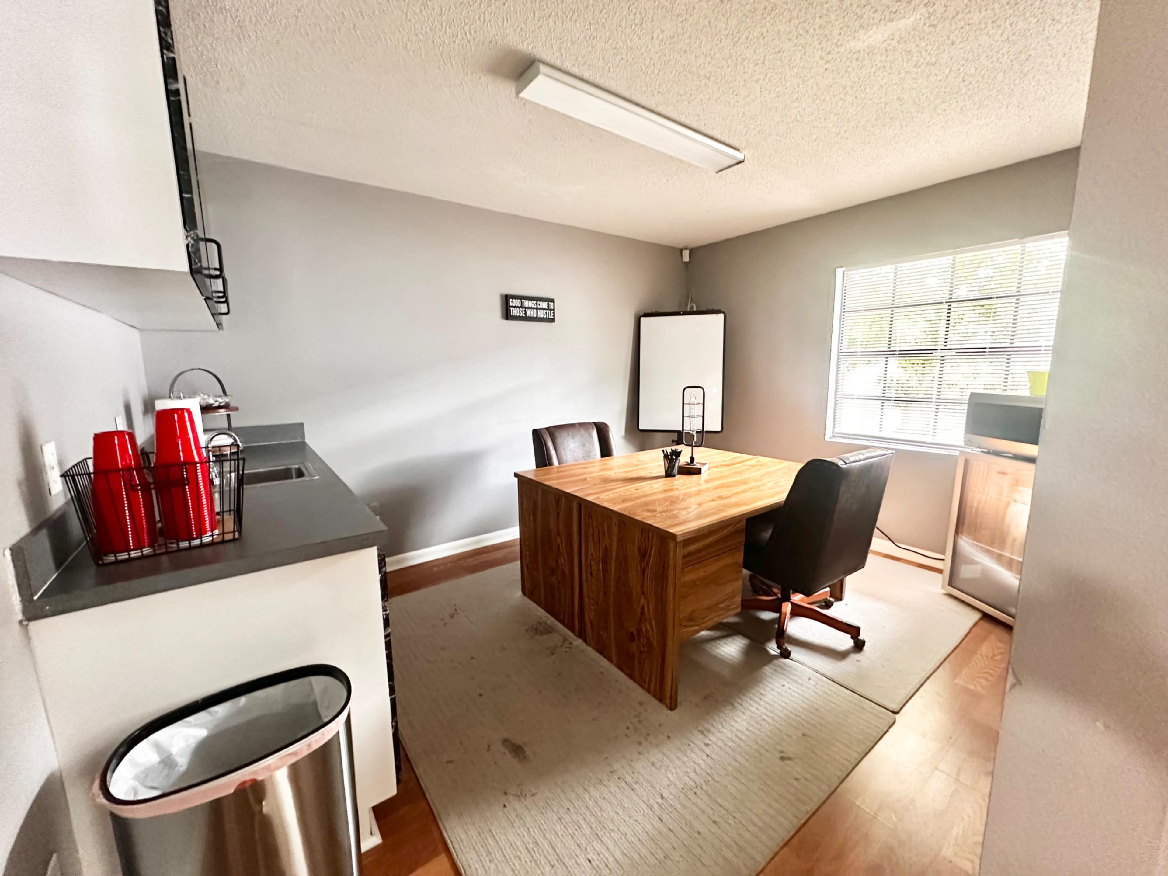 The image shows a modern office space featuring a wooden table with two chairs, a whiteboard, and kitchen amenities such as a refrigerator and trash can against the wall.