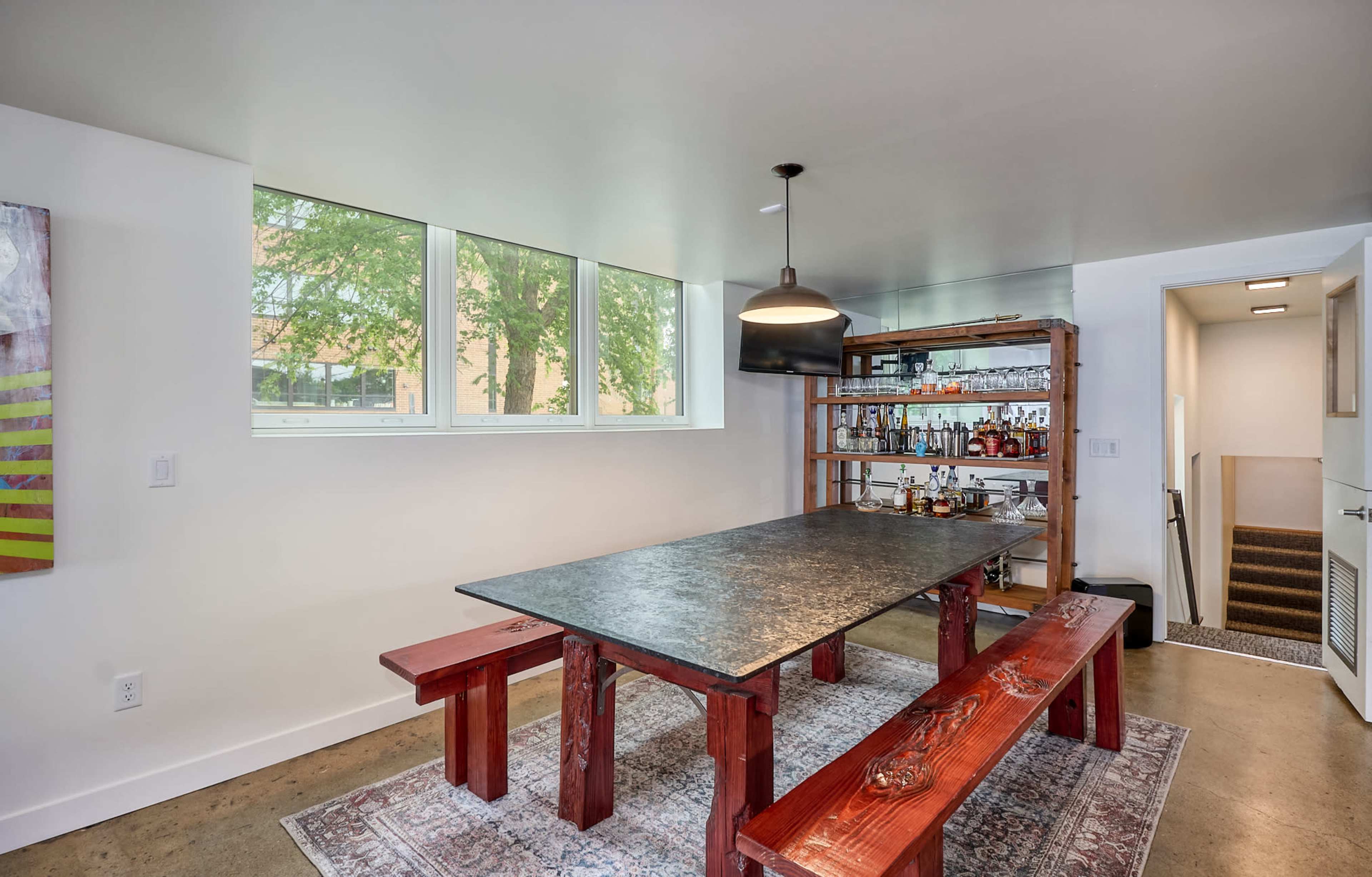 A modern dining area featuring a large black table with red wooden benches, a shelf with bottles in the background, and windows allowing natural light to enter.