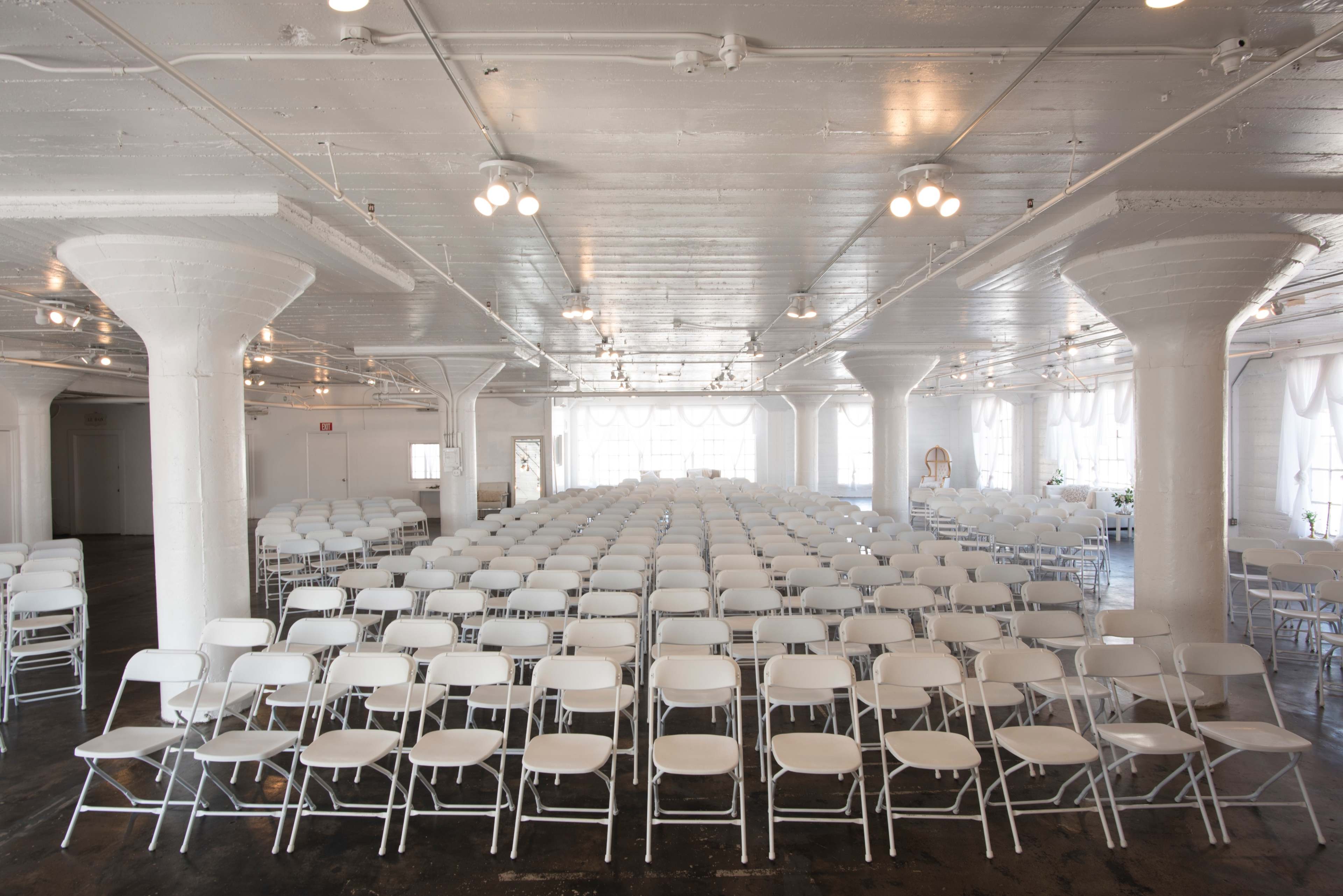 The image shows a large, brightly lit room filled with rows of empty white folding chairs arranged for an event.