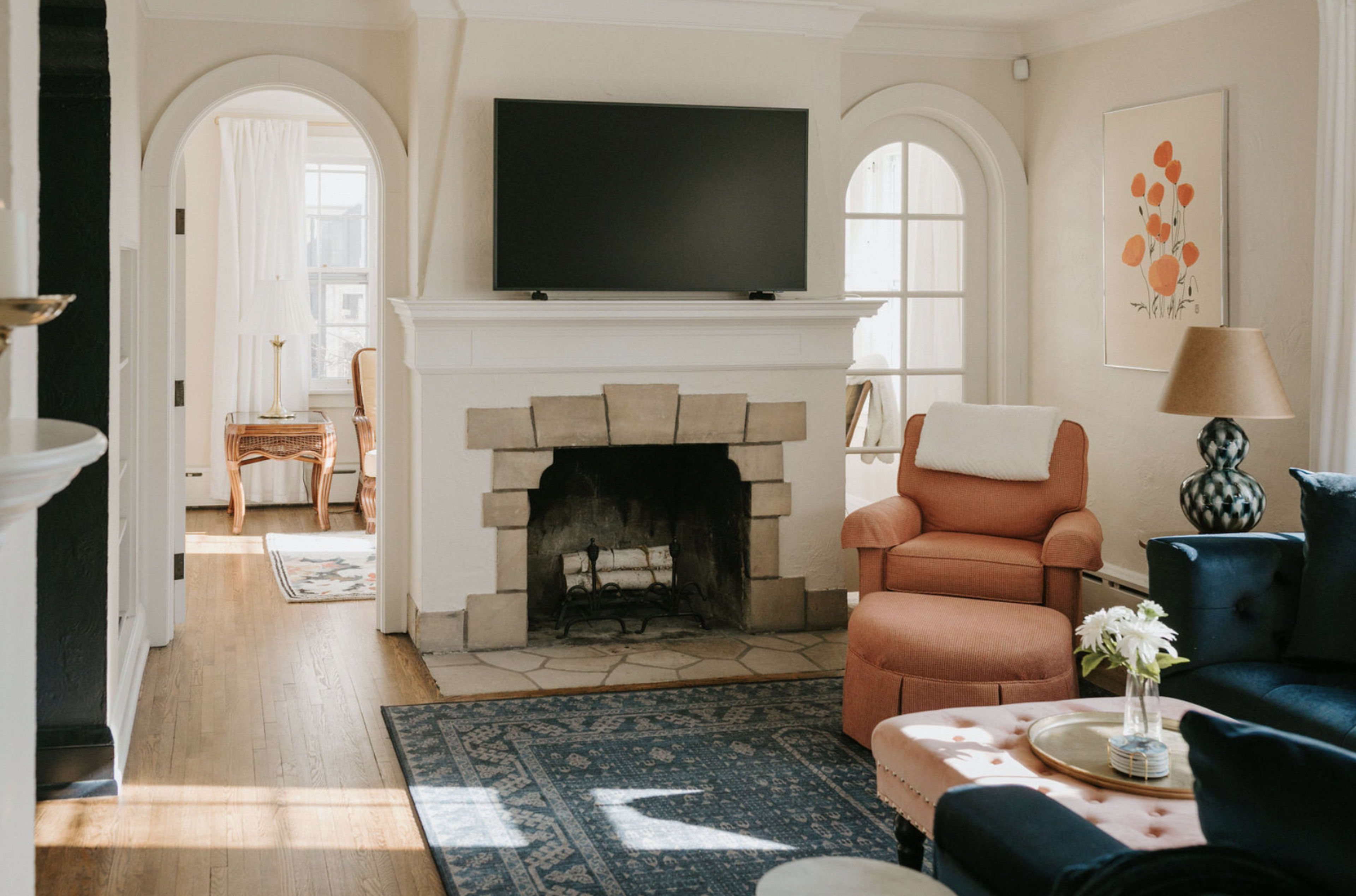 The image shows a cozy living room featuring a stone fireplace, a television mounted above it, a pink armchair, and a navy blue couch, with light streaming in from the adjoining room.