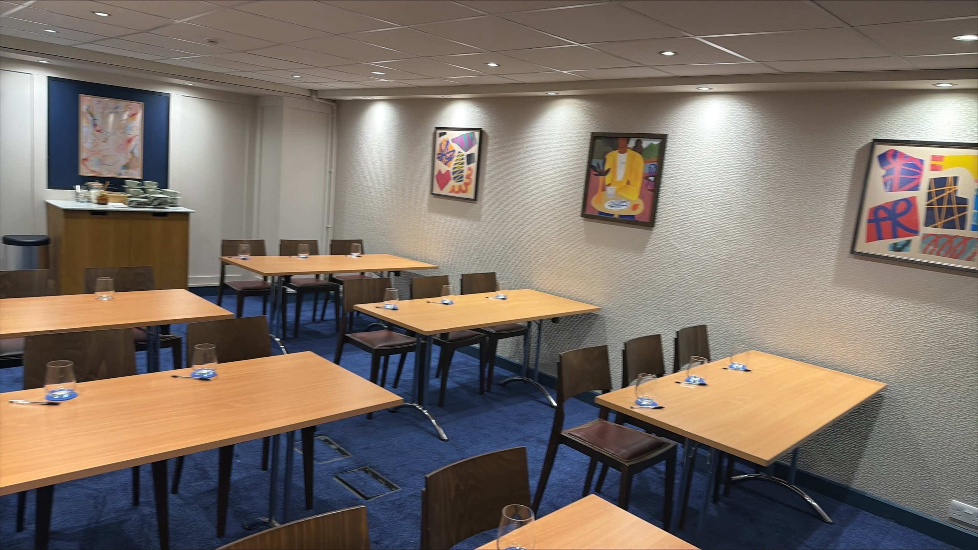 The image shows a meeting room equipped with several wooden tables and chairs, along with colorful artwork on the walls.