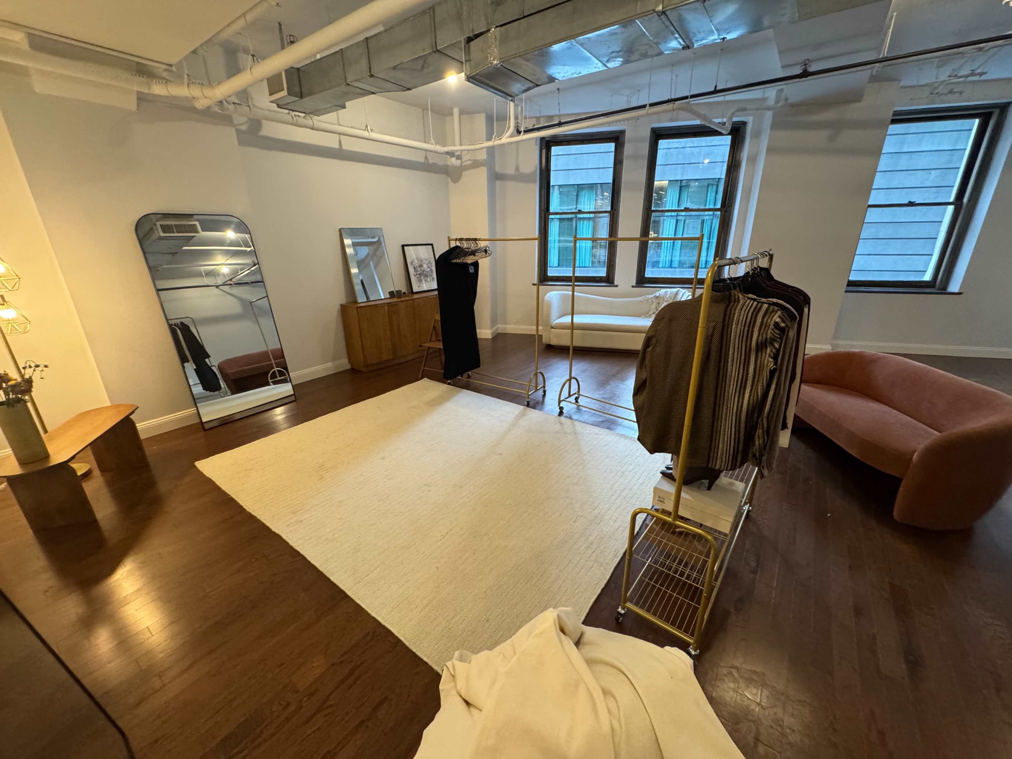 The image shows a room with hardwood floors featuring a large mirror, a clothing rack with garments, a stylish couch, and a minimalist design aesthetic.