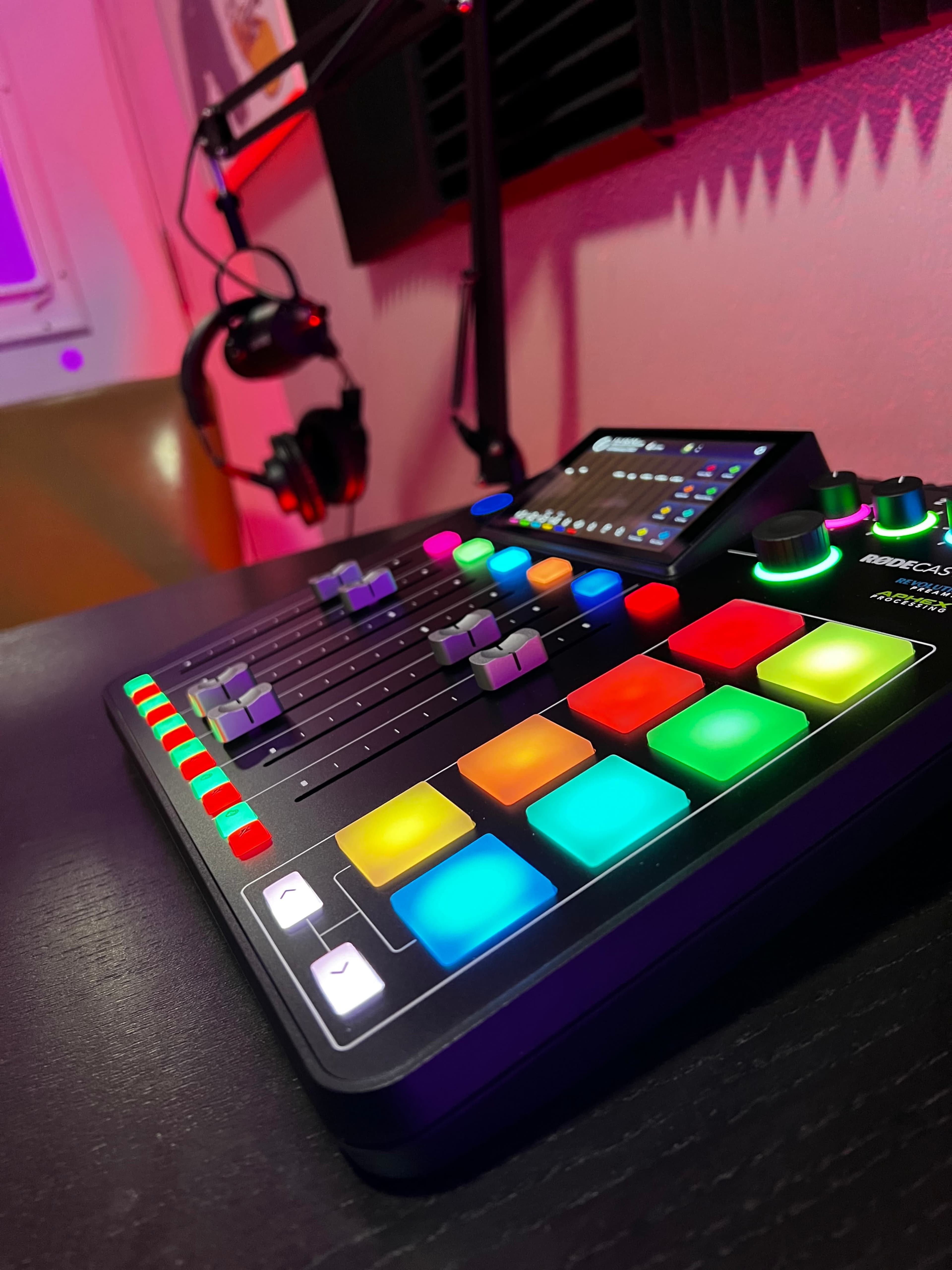 A colorful audio mixer with several sliders and buttons is positioned on a desk, alongside a pair of headphones in a dimly lit room.