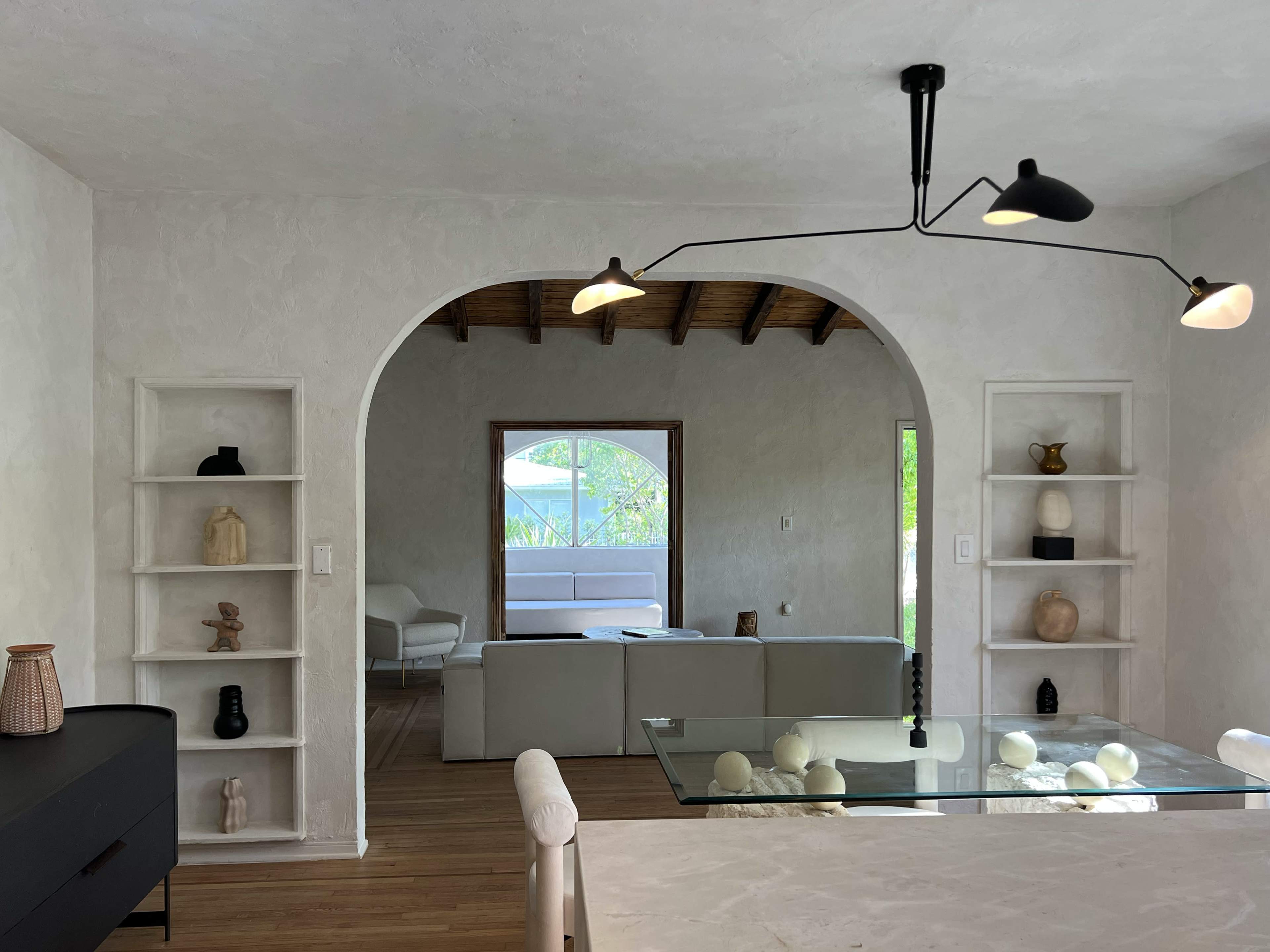 The image shows a living space with arched openings, featuring white walls, wooden ceilings, and shelves displaying decorative objects.