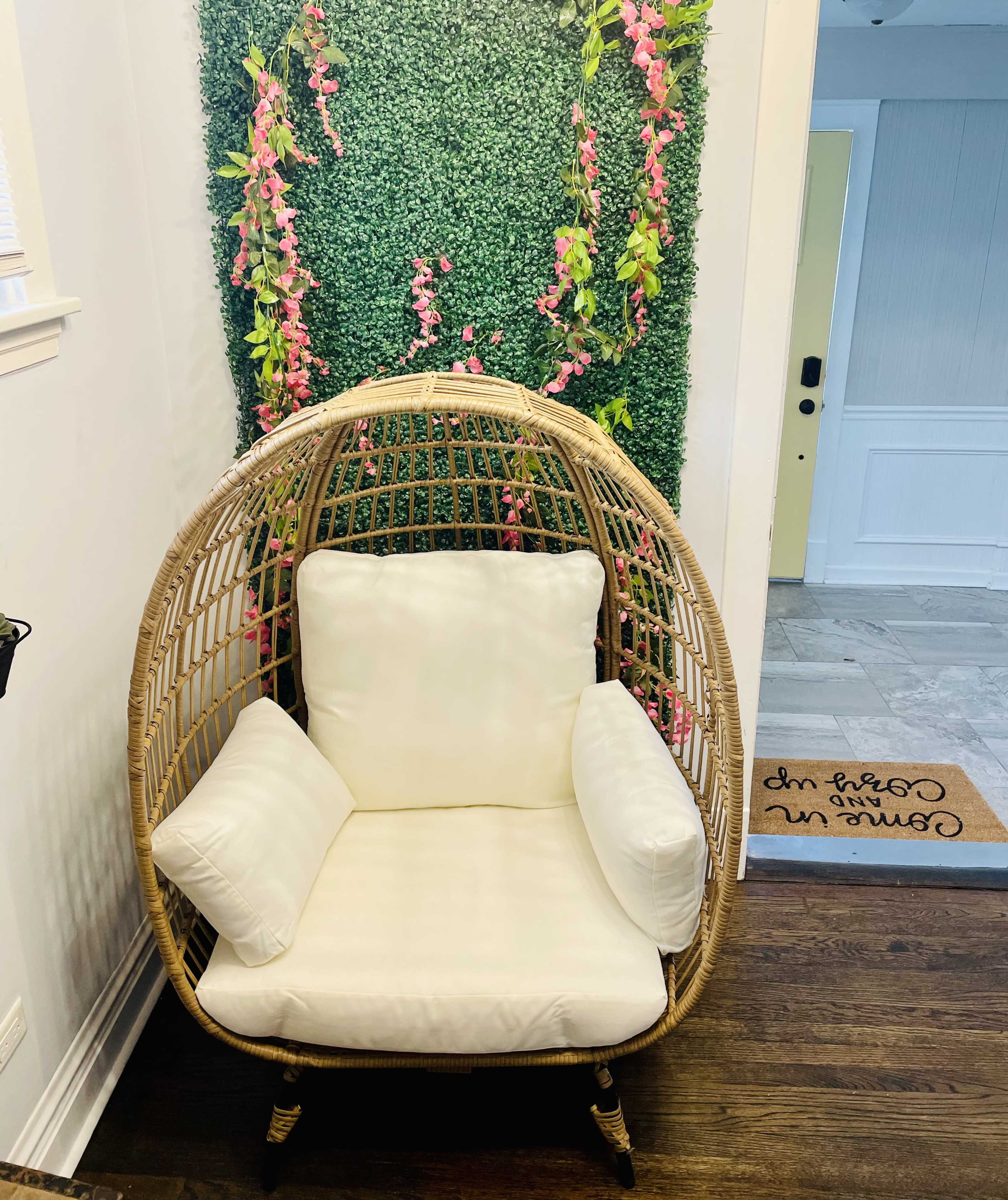 A hanging rattan chair with white cushions is positioned in front of a wall adorned with green foliage and pink flowers.