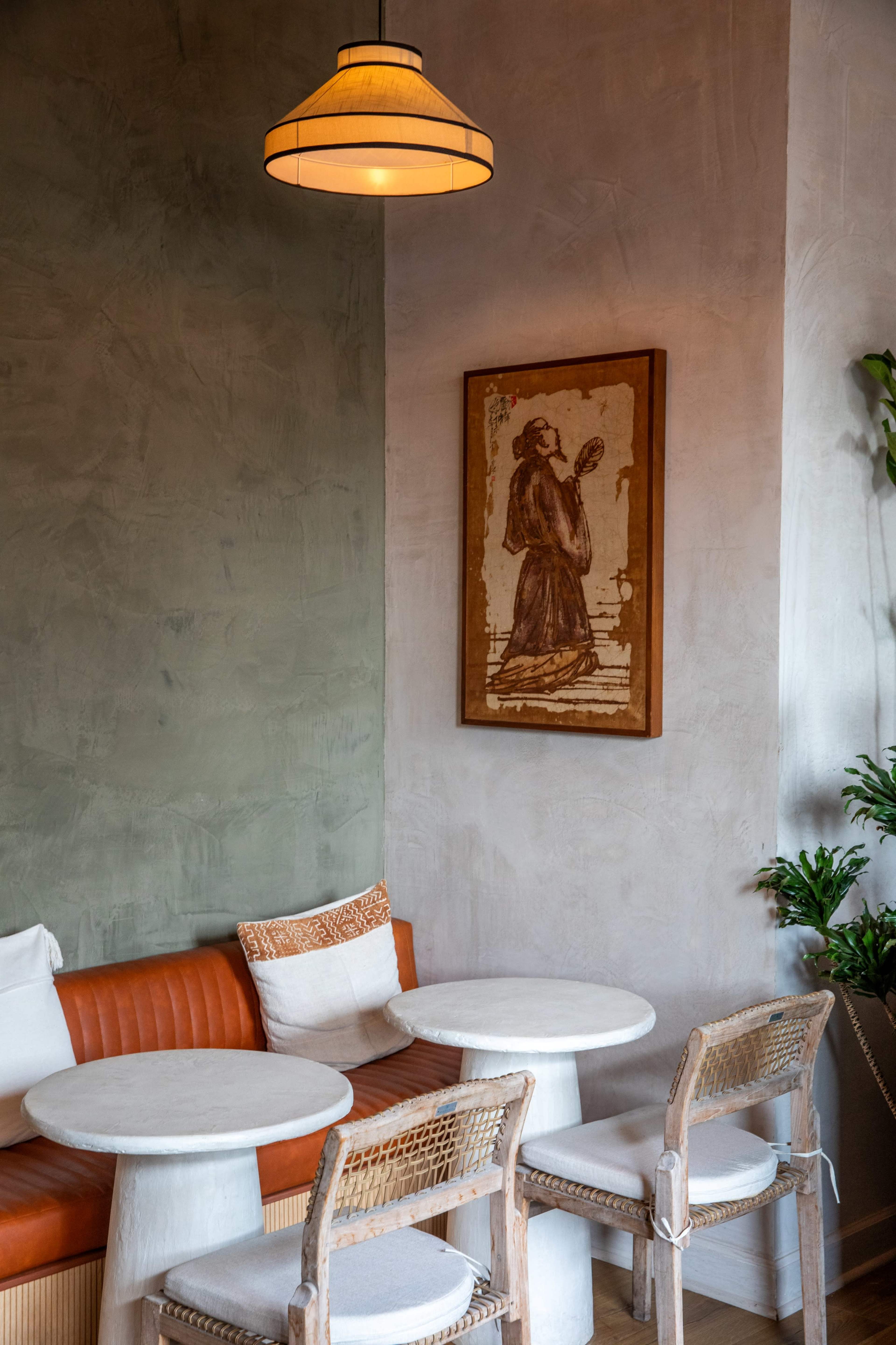 A cozy café corner features a leather seat, two round tables, wooden chairs, and a framed artwork on the wall.