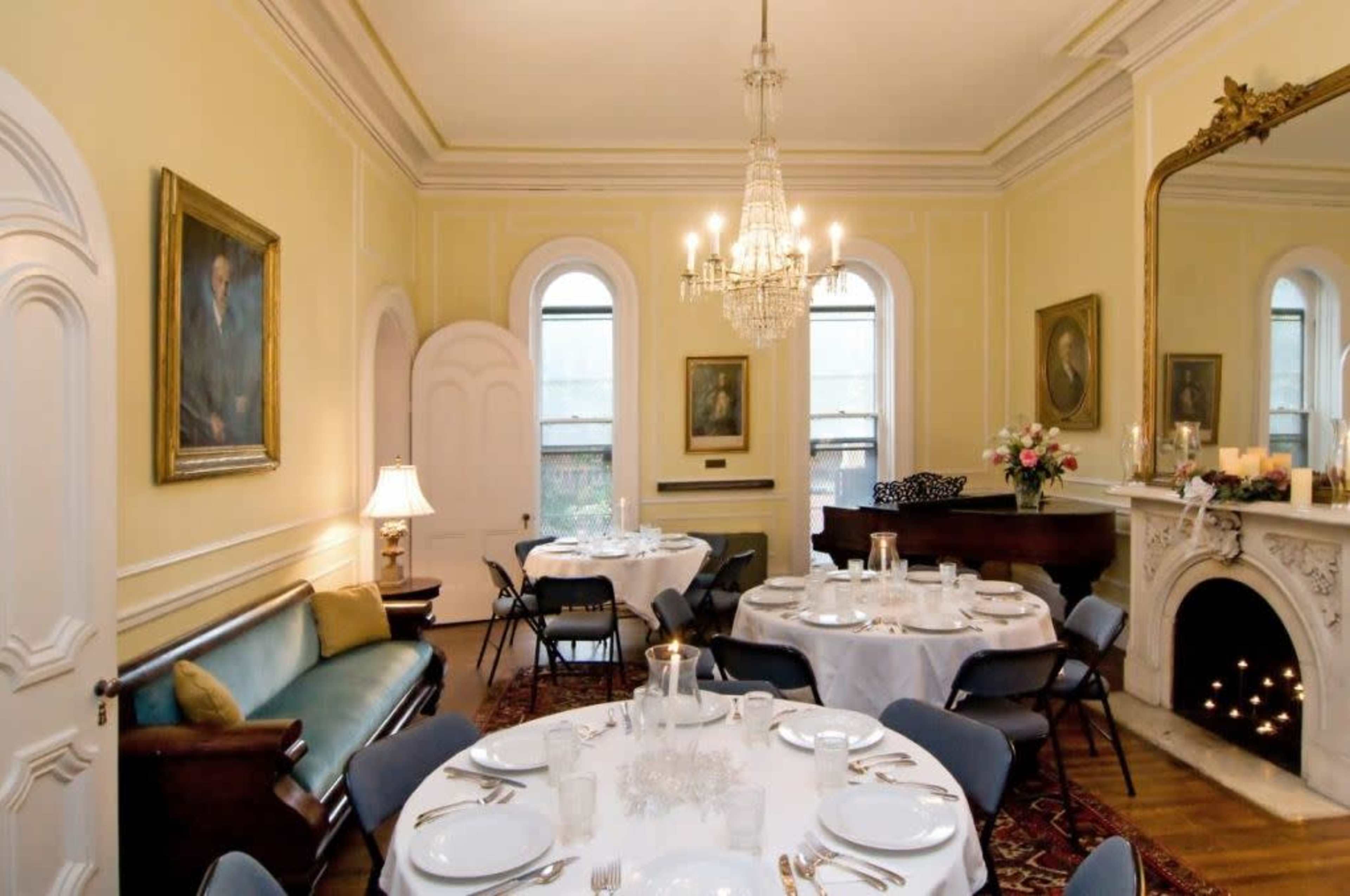 A dining room with a round table set for a meal, surrounded by chairs, and featuring a chandelier, a fireplace, and several framed paintings on the walls.