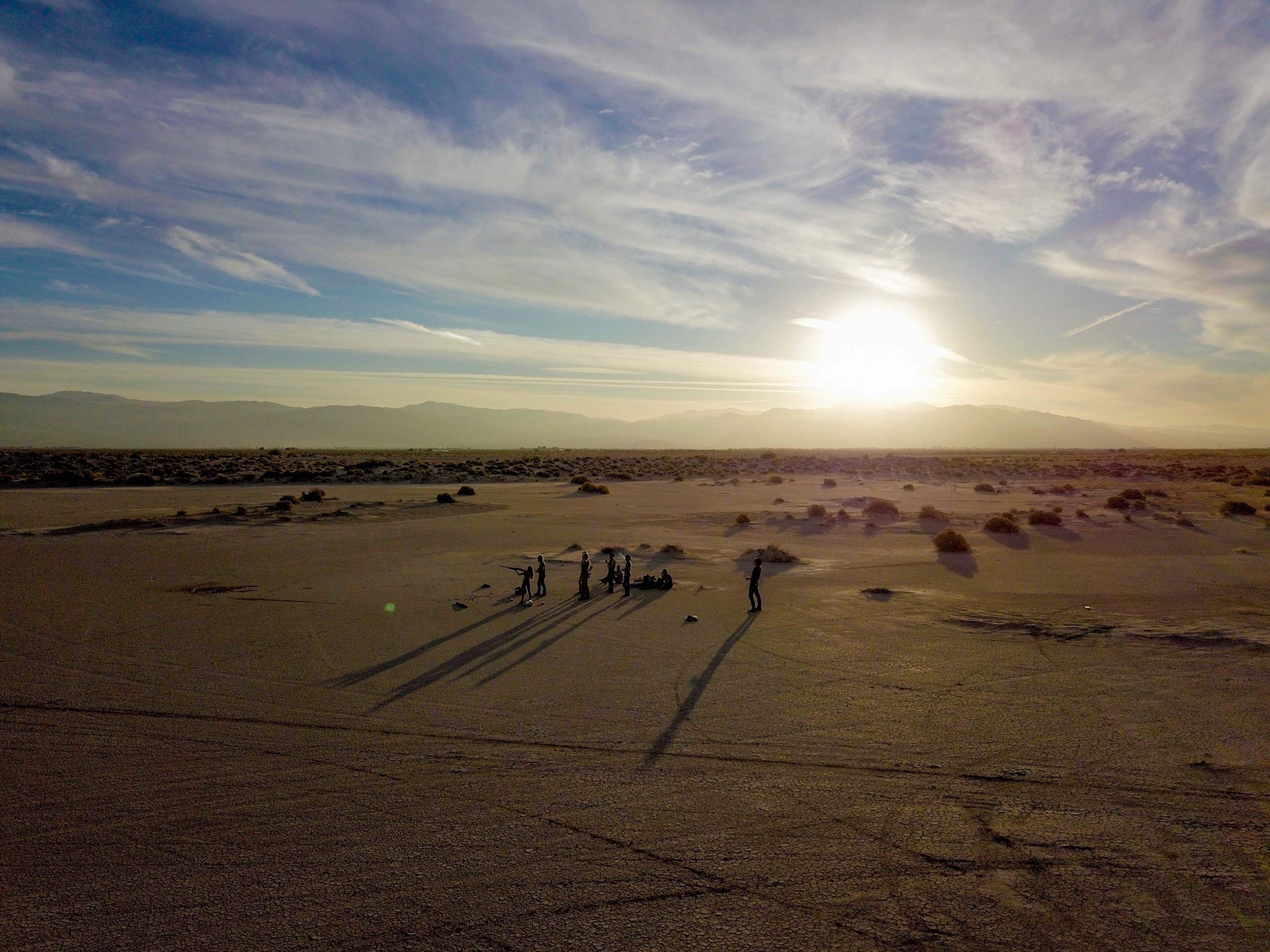 A group of people gathers in a desert landscape under a setting sun, casting long shadows on the sandy ground.