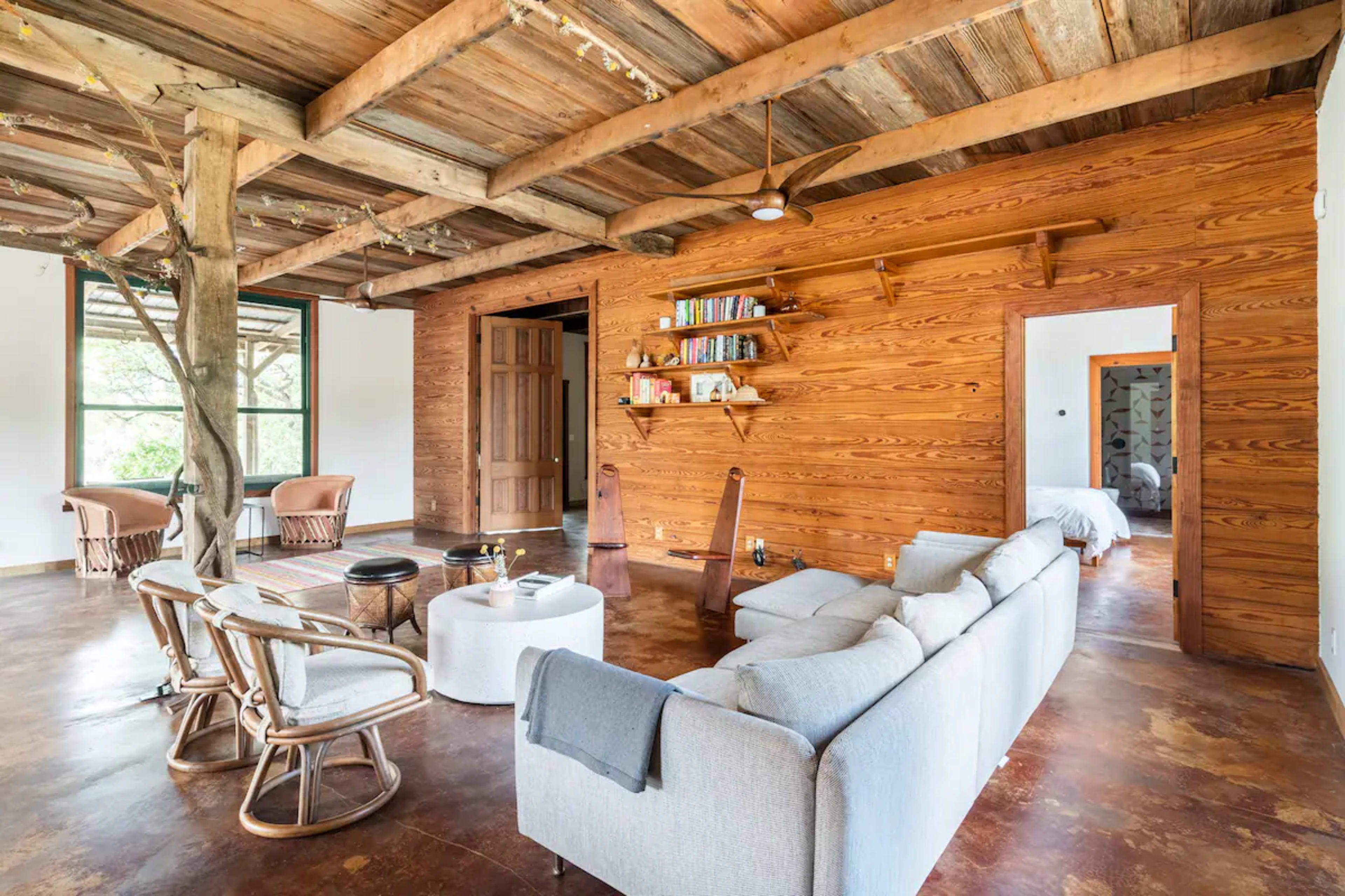 The image shows a rustic living room featuring wooden walls, a gray sectional sofa, a round coffee table, and various seating arrangements, with a doorway leading to another room.