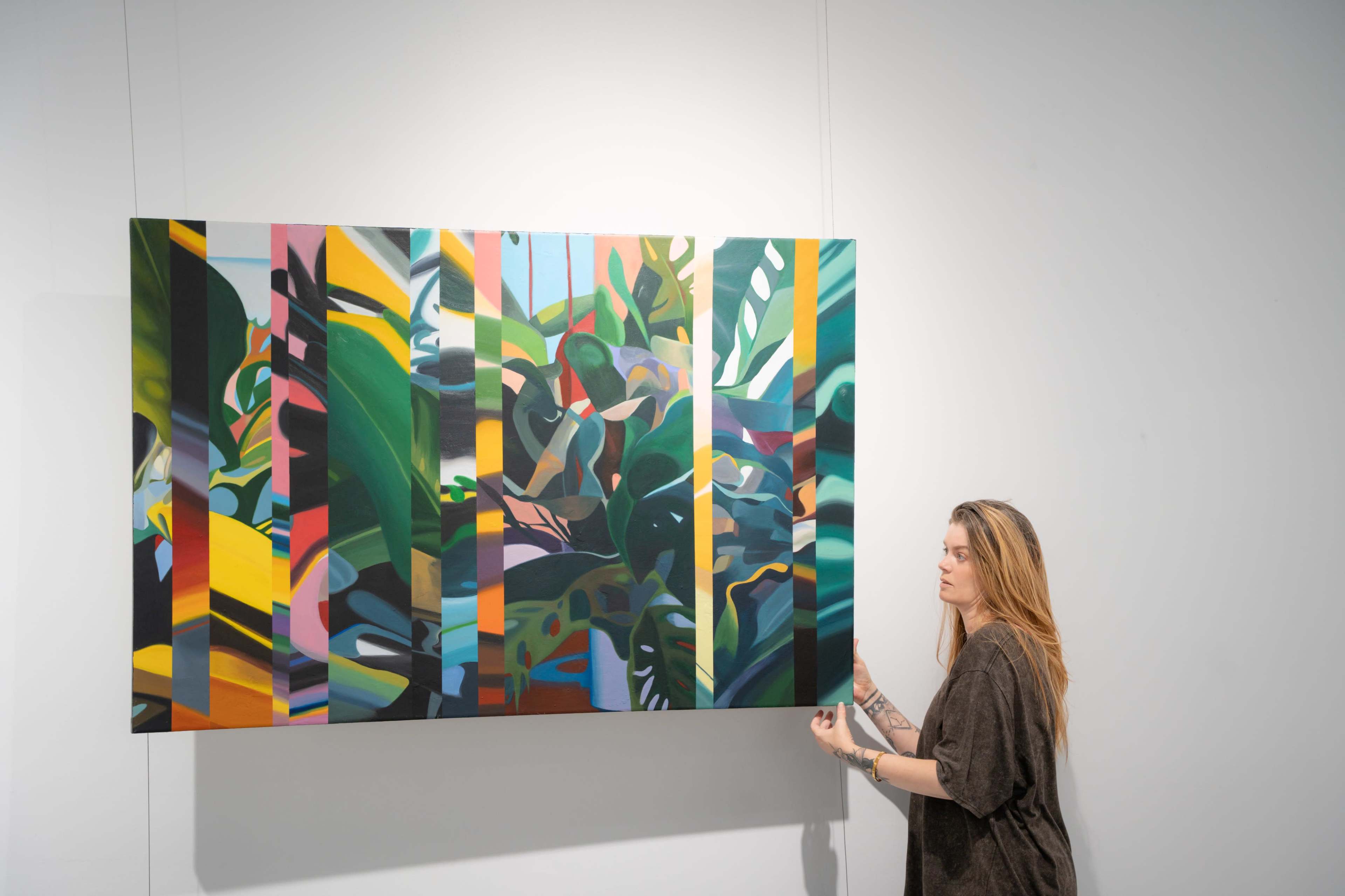A person is adjusting a colorful abstract painting of tropical foliage hung on a white wall.