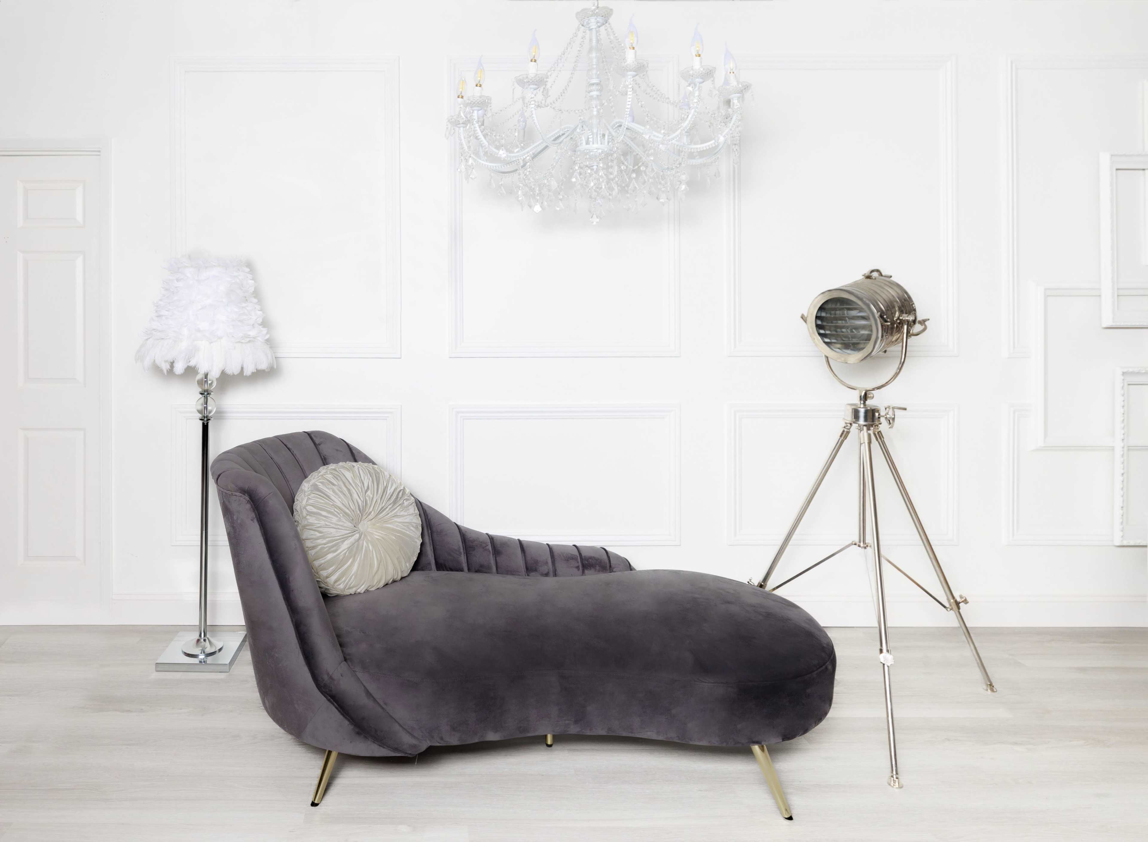 A modern gray chaise lounge positioned against a white wall, accompanied by a decorative chandelier overhead and a vintage spotlight on a tripod nearby.