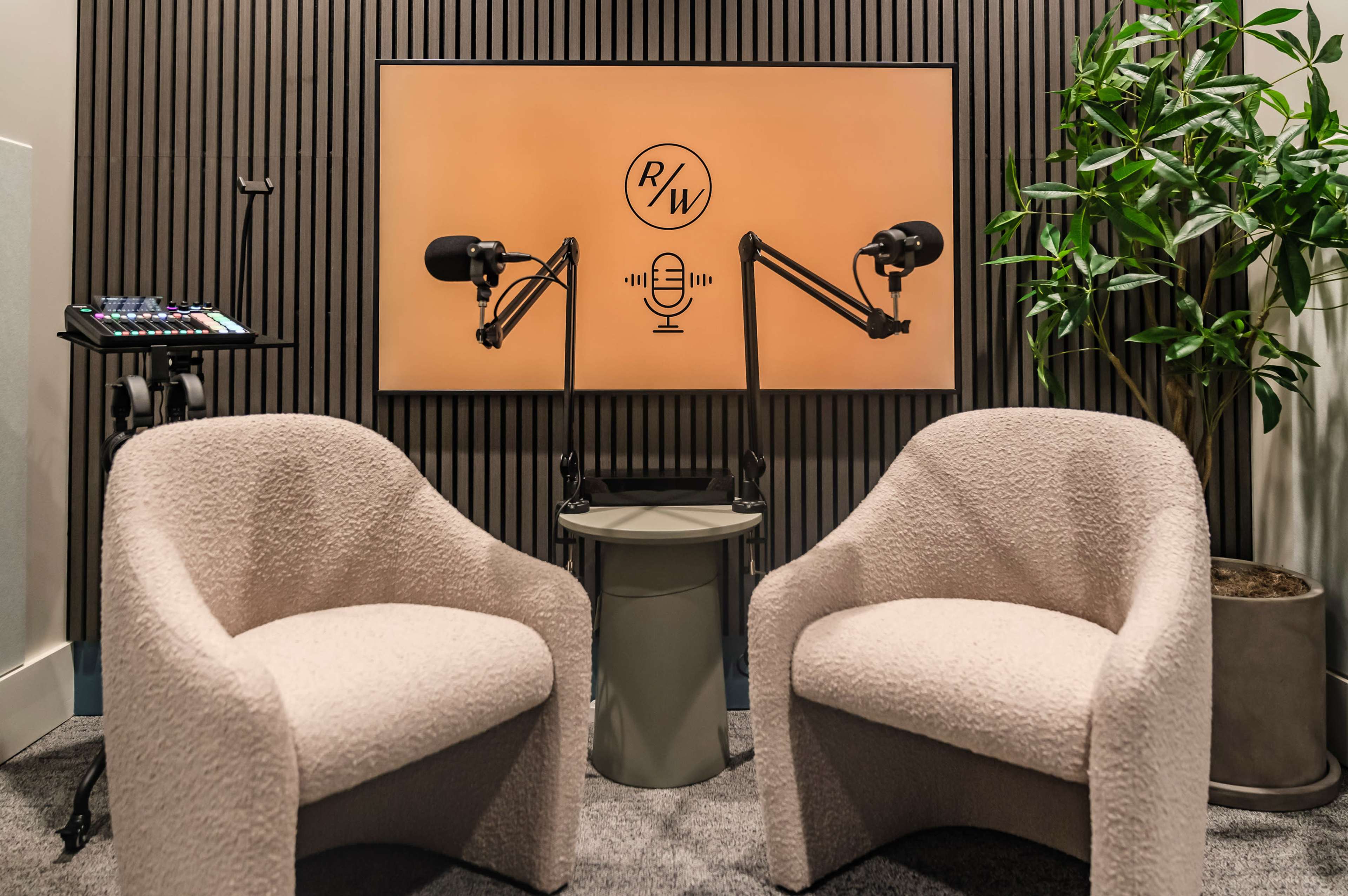 The image shows a podcast studio setup with two plush chairs facing a microphone stand and a wall-mounted screen featuring a logo.