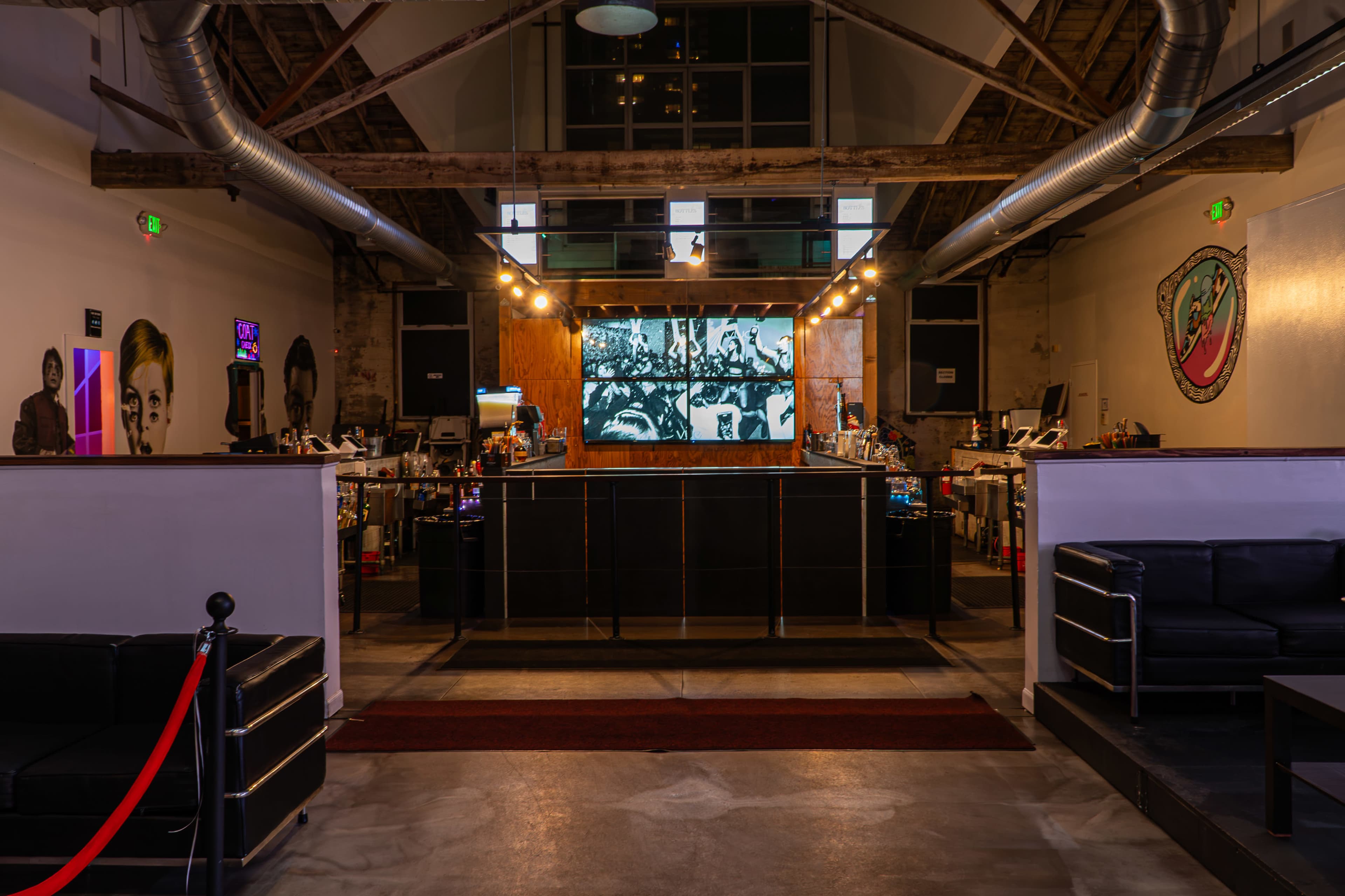 A modern interior space with a bar area and large screen displaying black-and-white images, surrounded by seating areas and industrial decor.