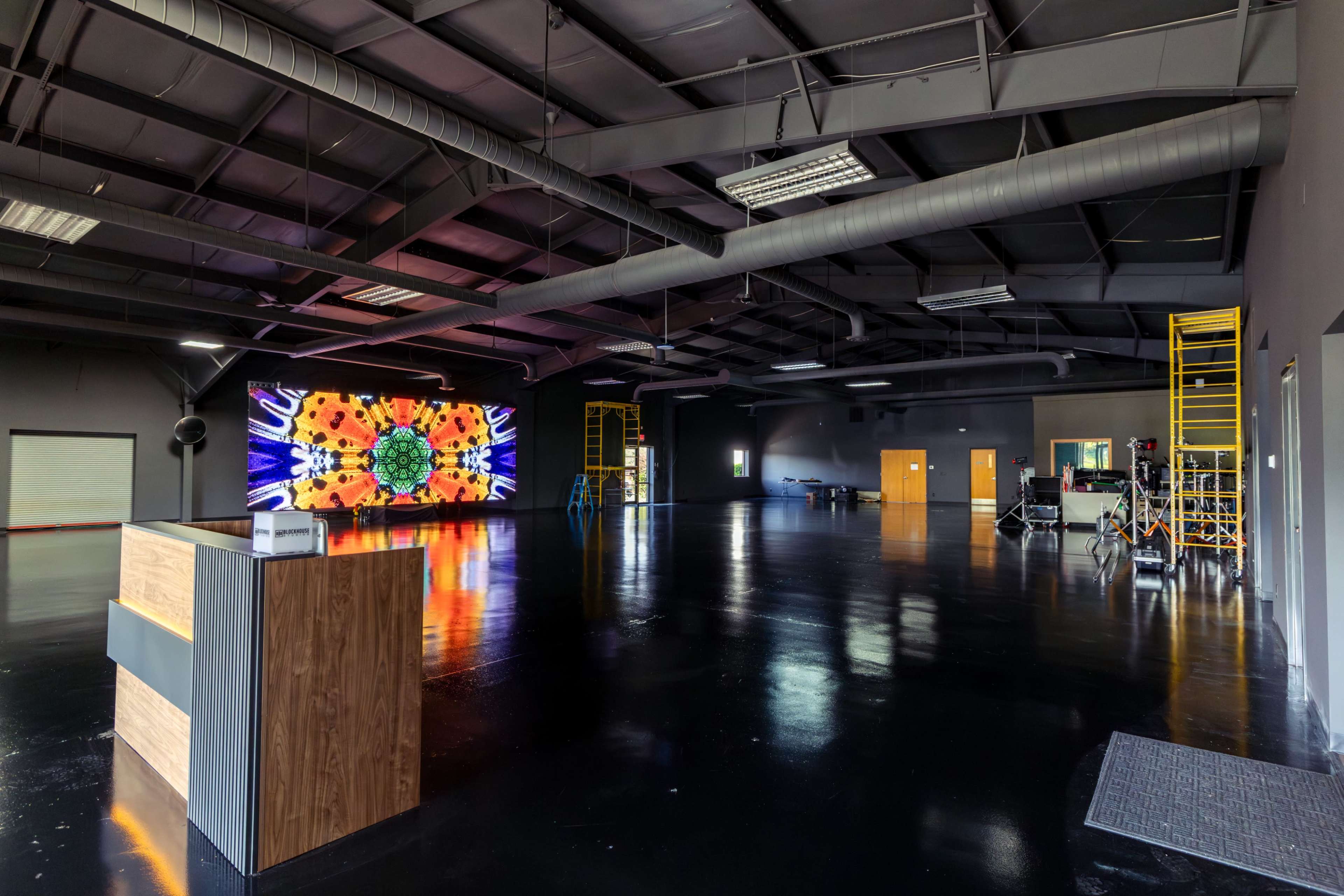 The image shows a large, open industrial space with a reception desk in the foreground and a colorful digital screen on the back wall.