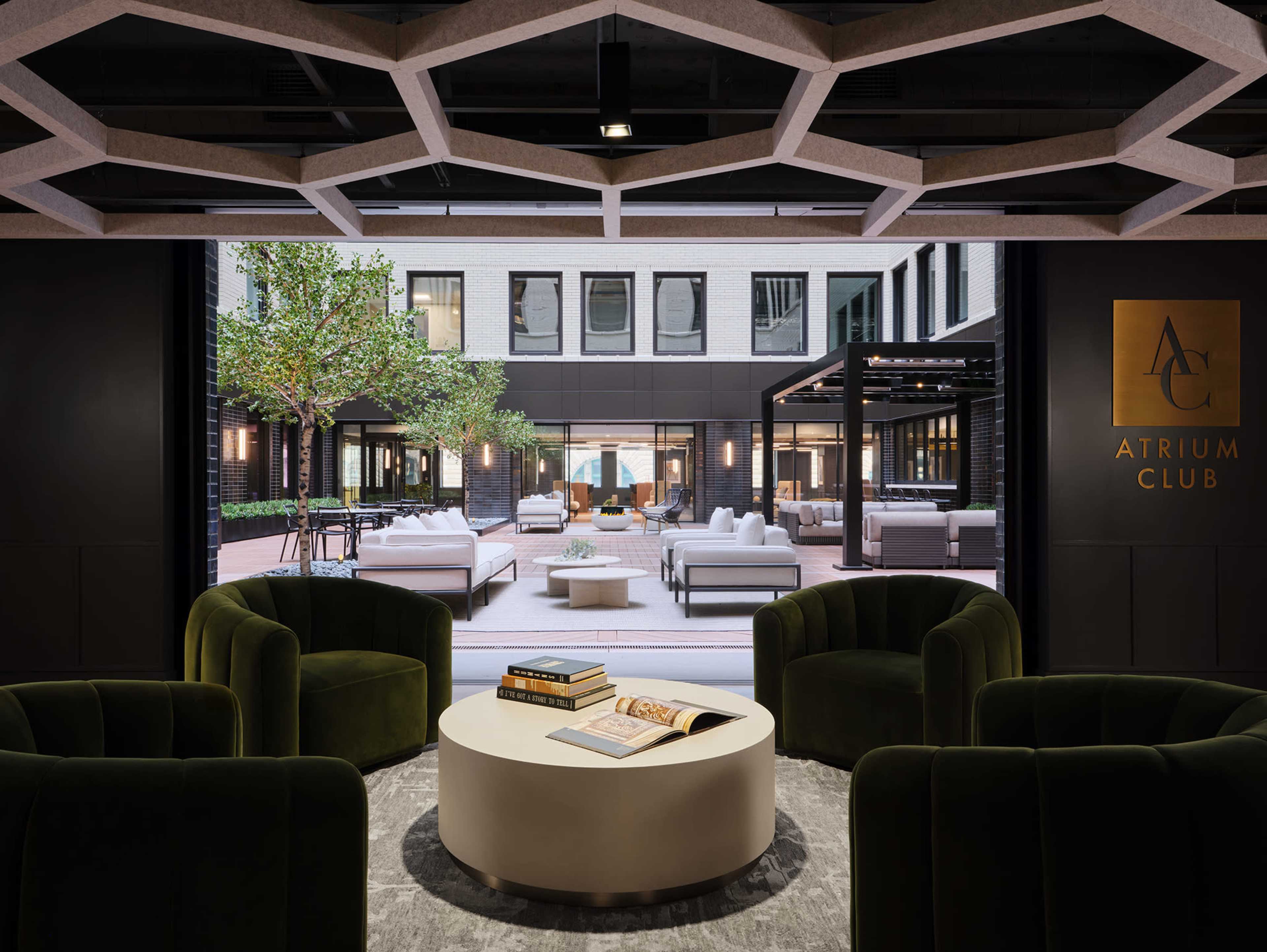 The image shows a modern lounge area with green armchairs and a round table, featuring a large window displaying an outdoor courtyard with seating and greenery.