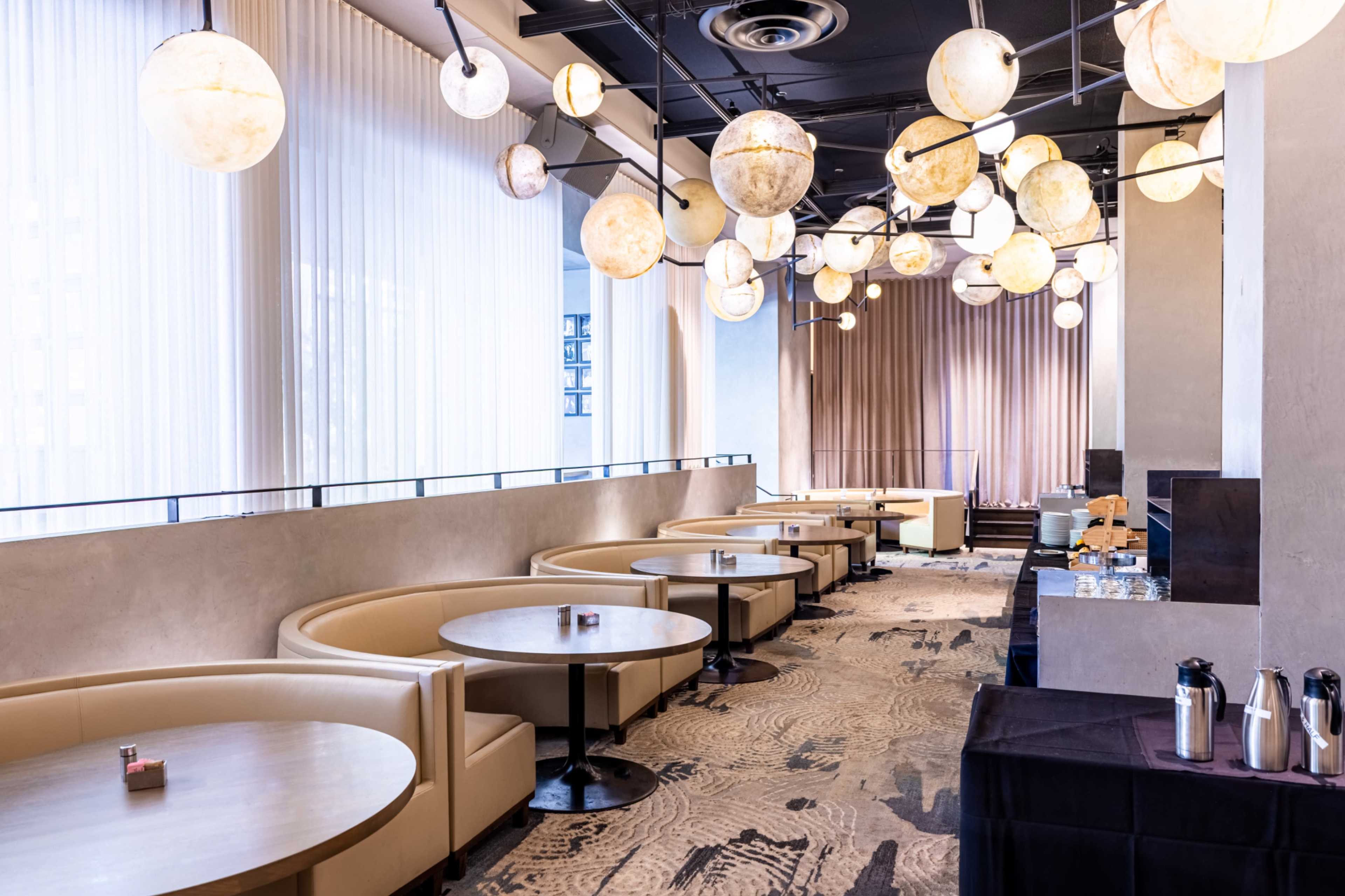 The image shows a modern restaurant interior featuring beige booth seating, round tables, and decorative light fixtures overhead.