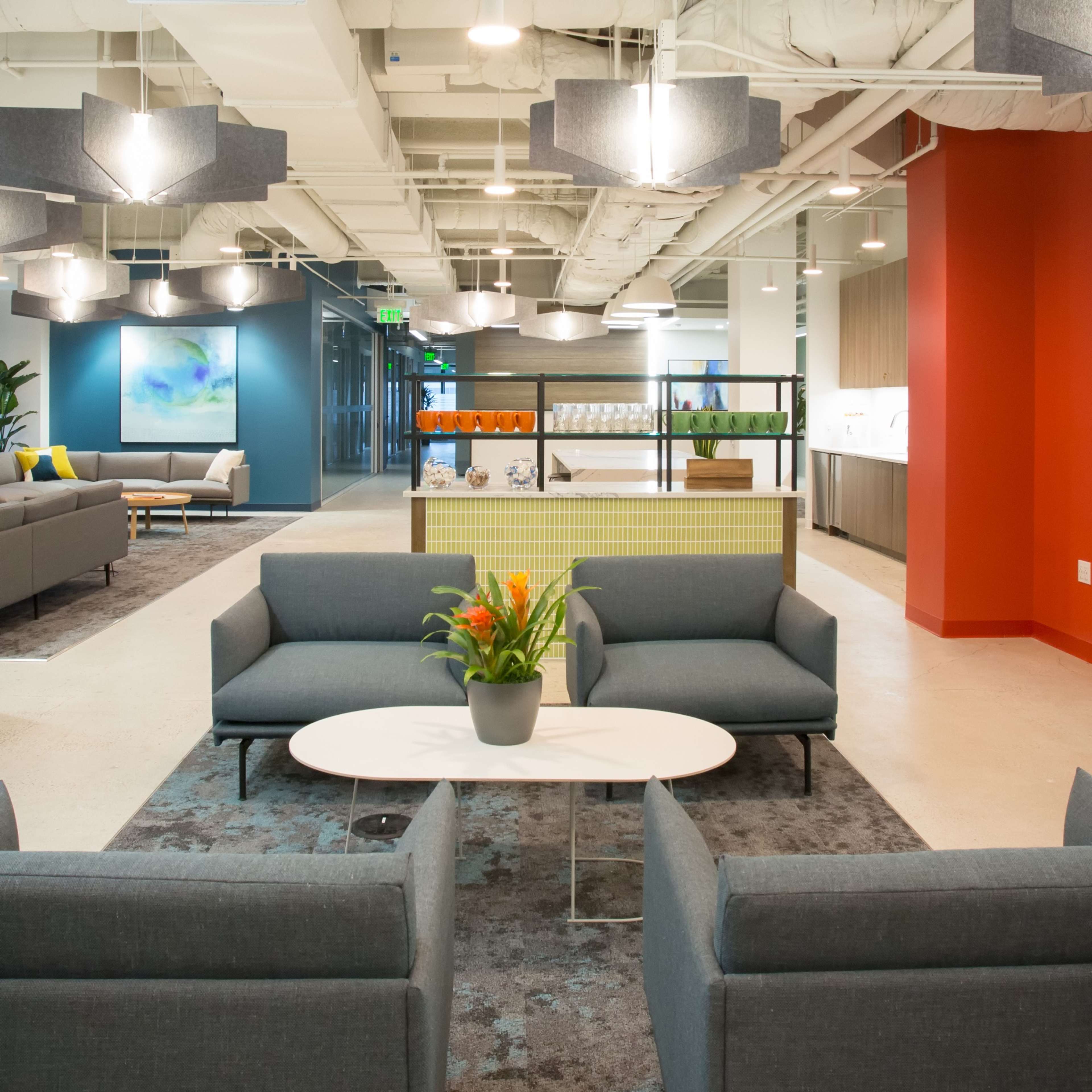 The image shows a modern office lounge area with gray sofas, a round coffee table, and pendant lighting, alongside a reception desk in a brightly colored section.