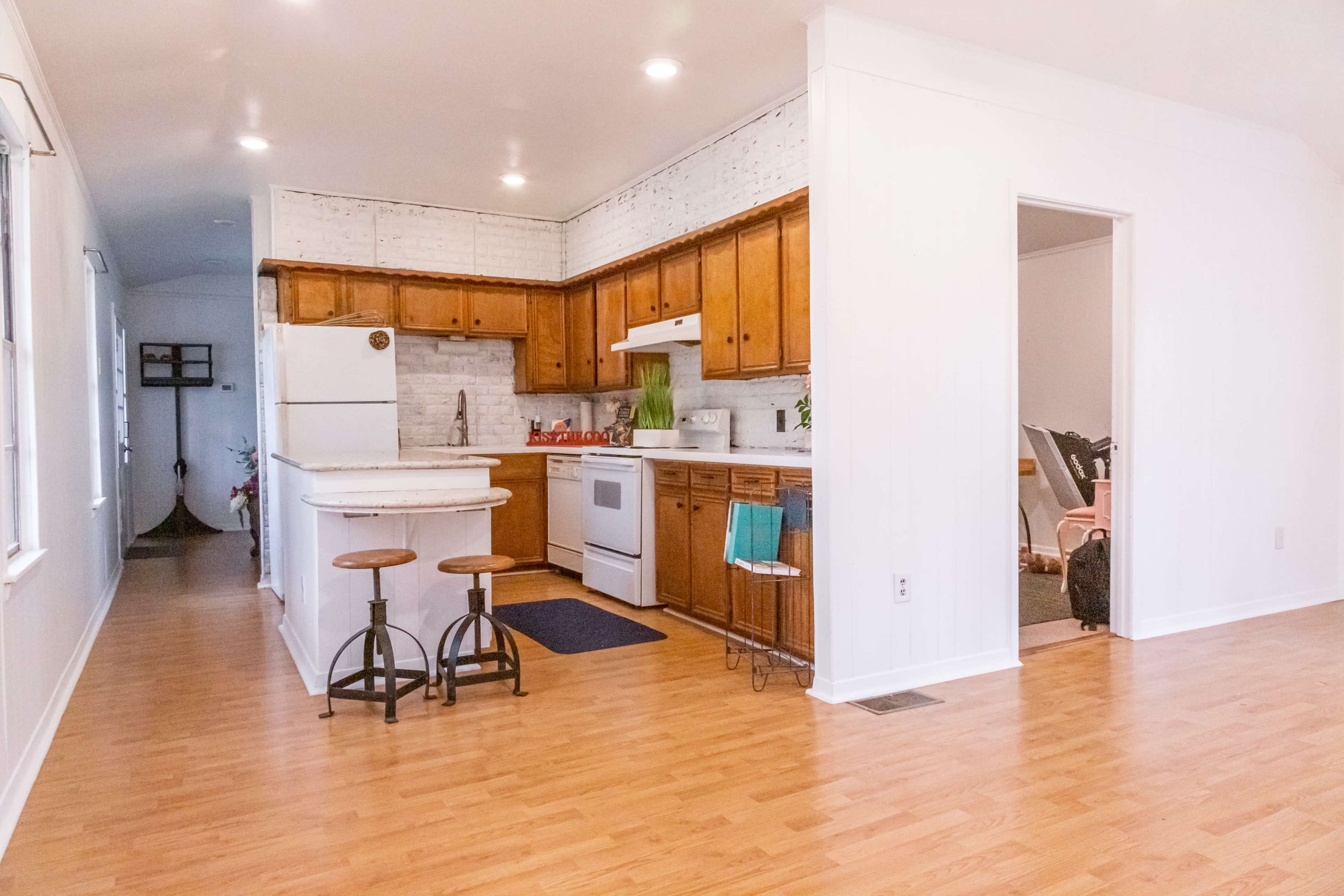 A kitchen area with wooden cabinets, a white refrigerator, and a small island with two stools, adjoining a light-filled space decorated with plants and furniture.