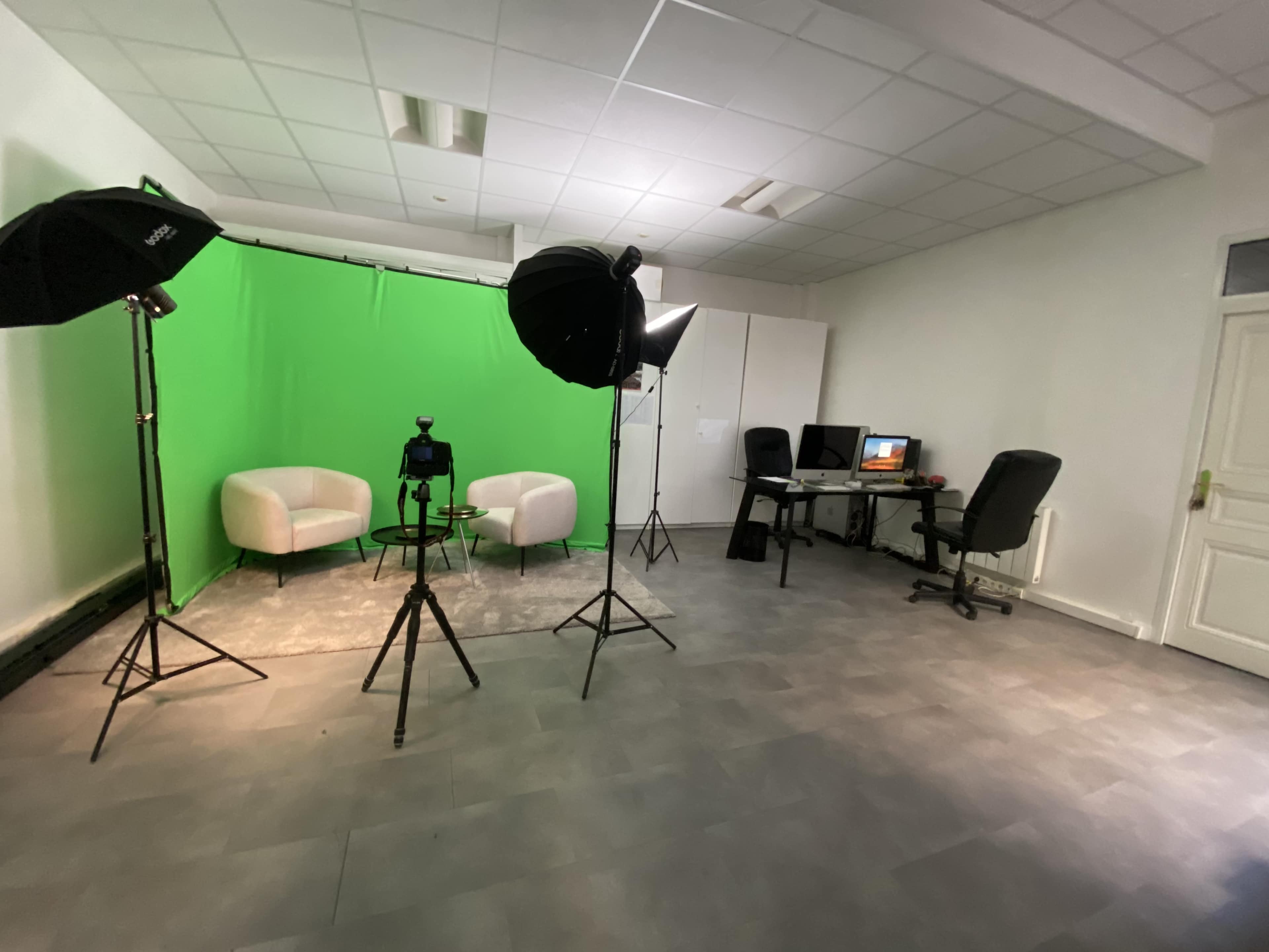 A studio setup with two white chairs, a green backdrop, lighting equipment, and a desk with a computer in a well-lit room.