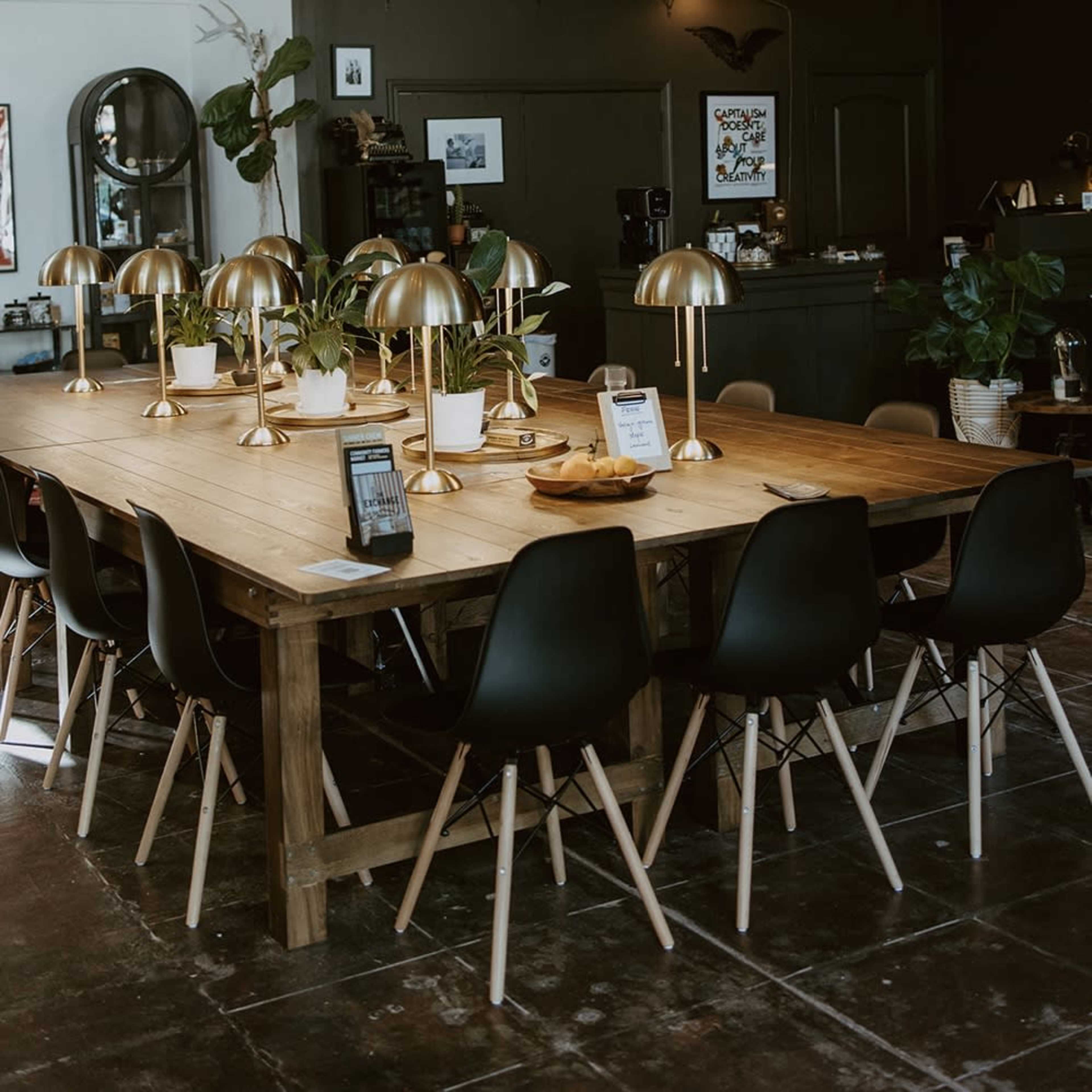 A large wooden dining table surrounded by black chairs and adorned with golden lamps and potted plants is set in a modern indoor space.