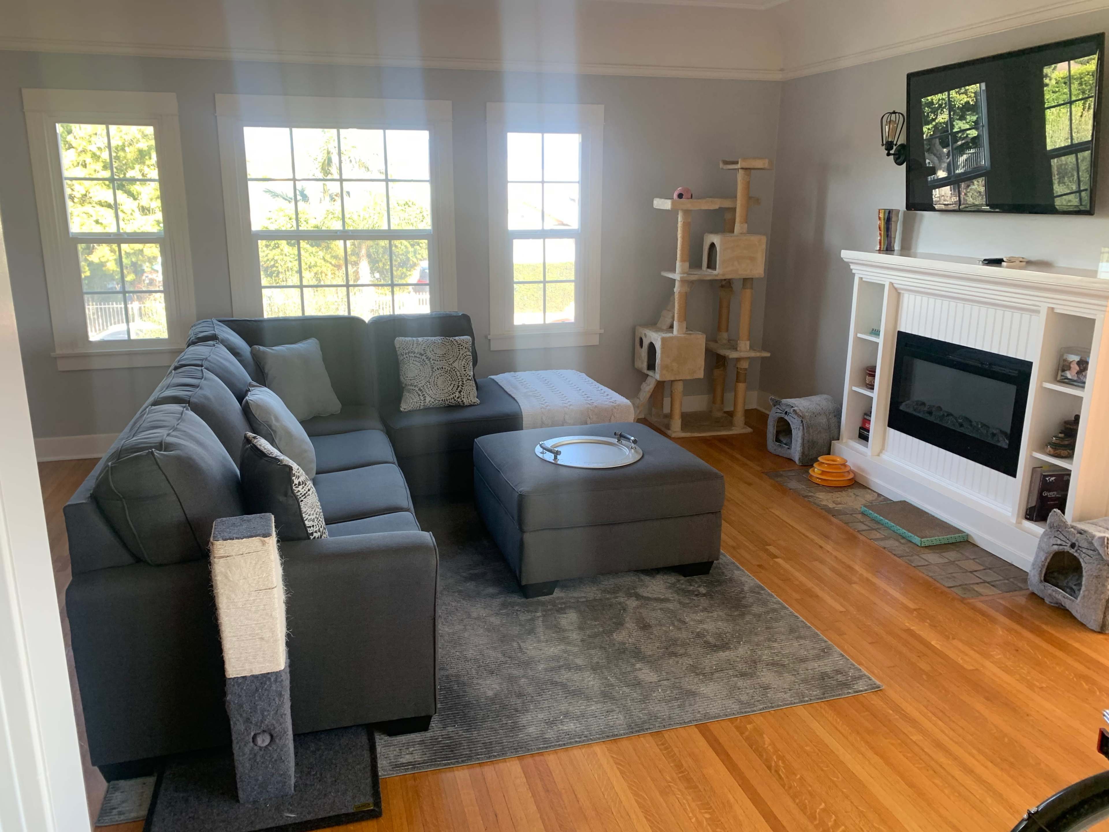 A cozy living room features a sectional couch, an ottoman, and a cat tree beside a fireplace with a television mounted above it.