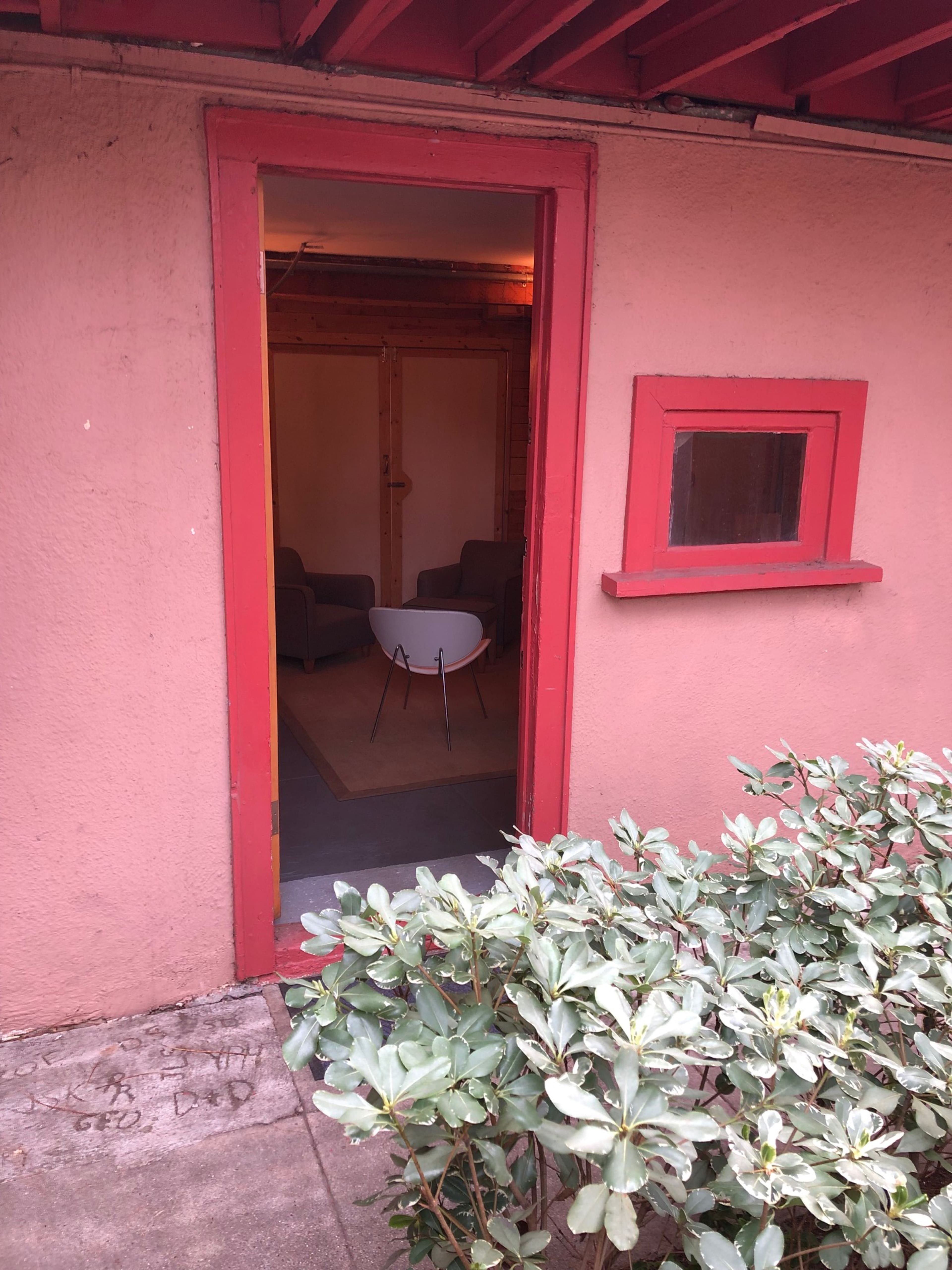 A short hallway leading to a small room furnished with two gray chairs and a white table, framed by a red doorway and surrounded by green shrubbery.