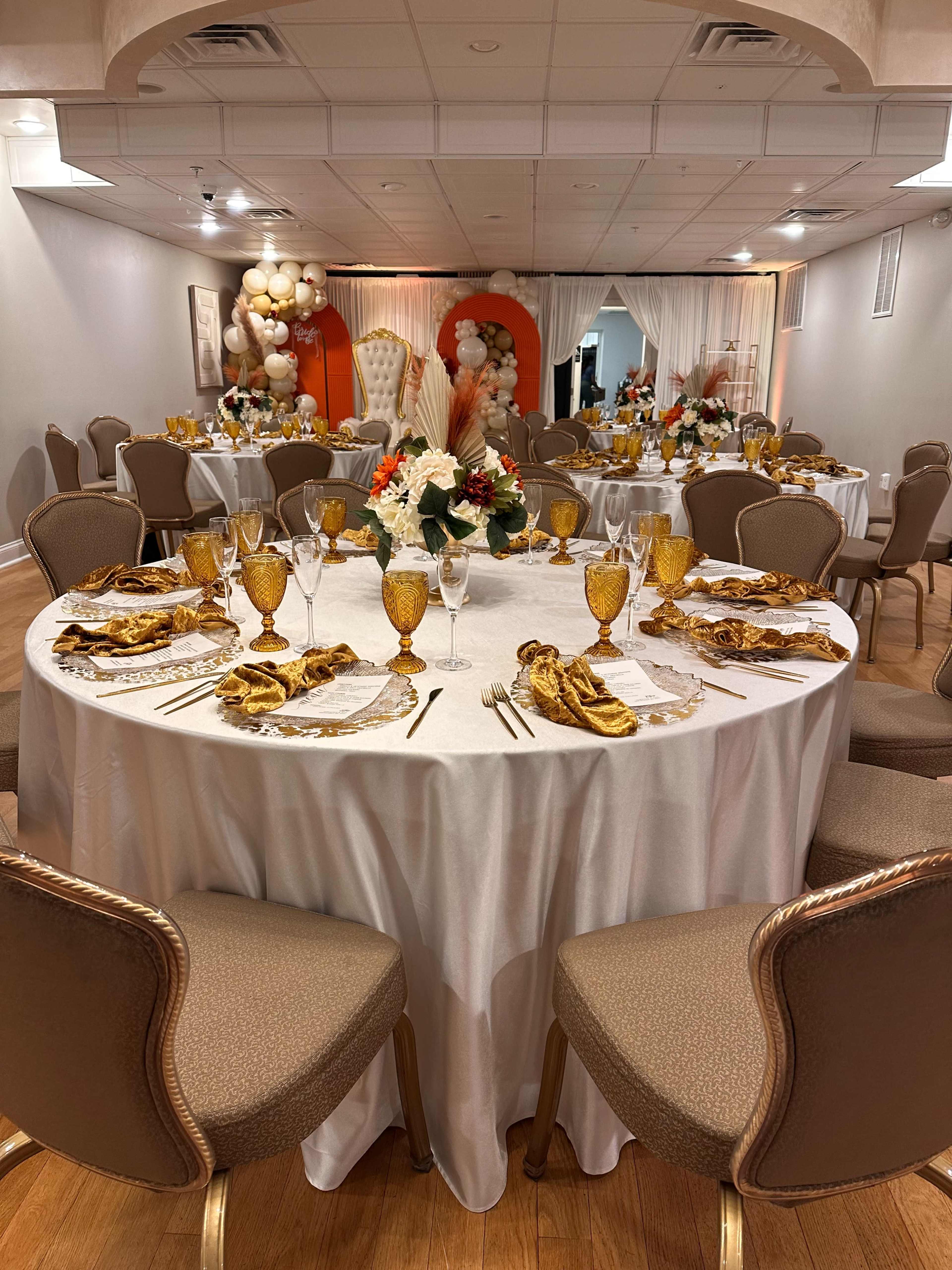 The image shows a decorated banquet hall with elegantly set round tables featuring white tablecloths, gold accents, and floral centerpieces.