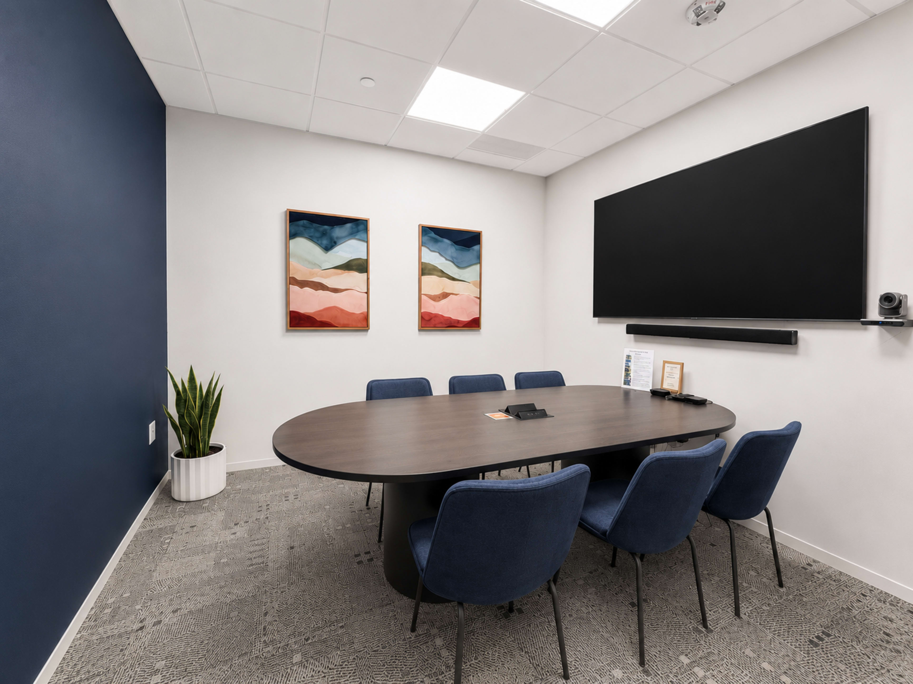 A modern conference room features an oval table surrounded by blue chairs, with two framed artwork pieces on the wall and a flat-screen monitor mounted above a shelf.