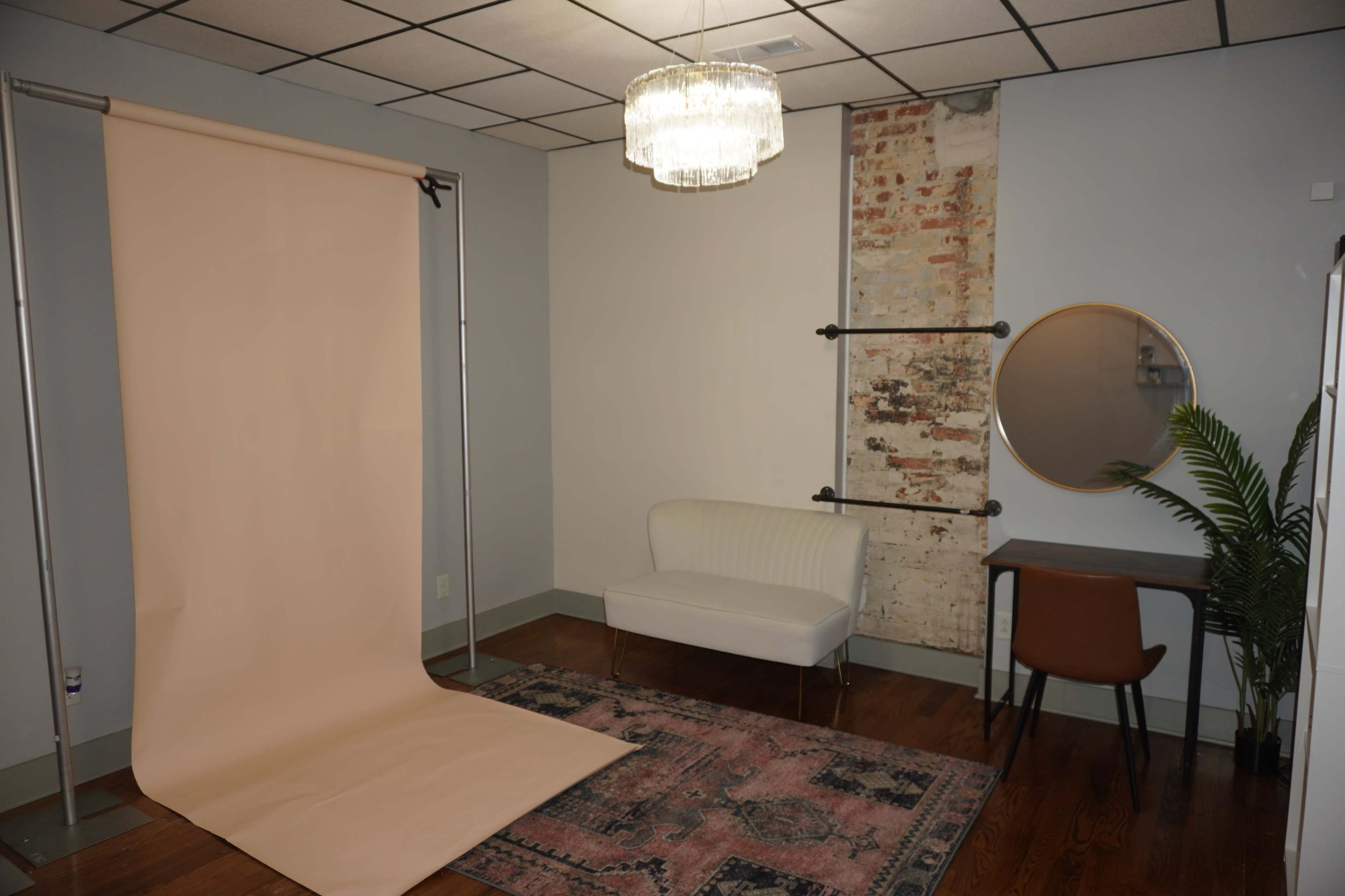 The image shows a photography studio with a soft pink backdrop, a white couch, a round mirror, a wooden desk, and a chandeliers hanging from the ceiling.