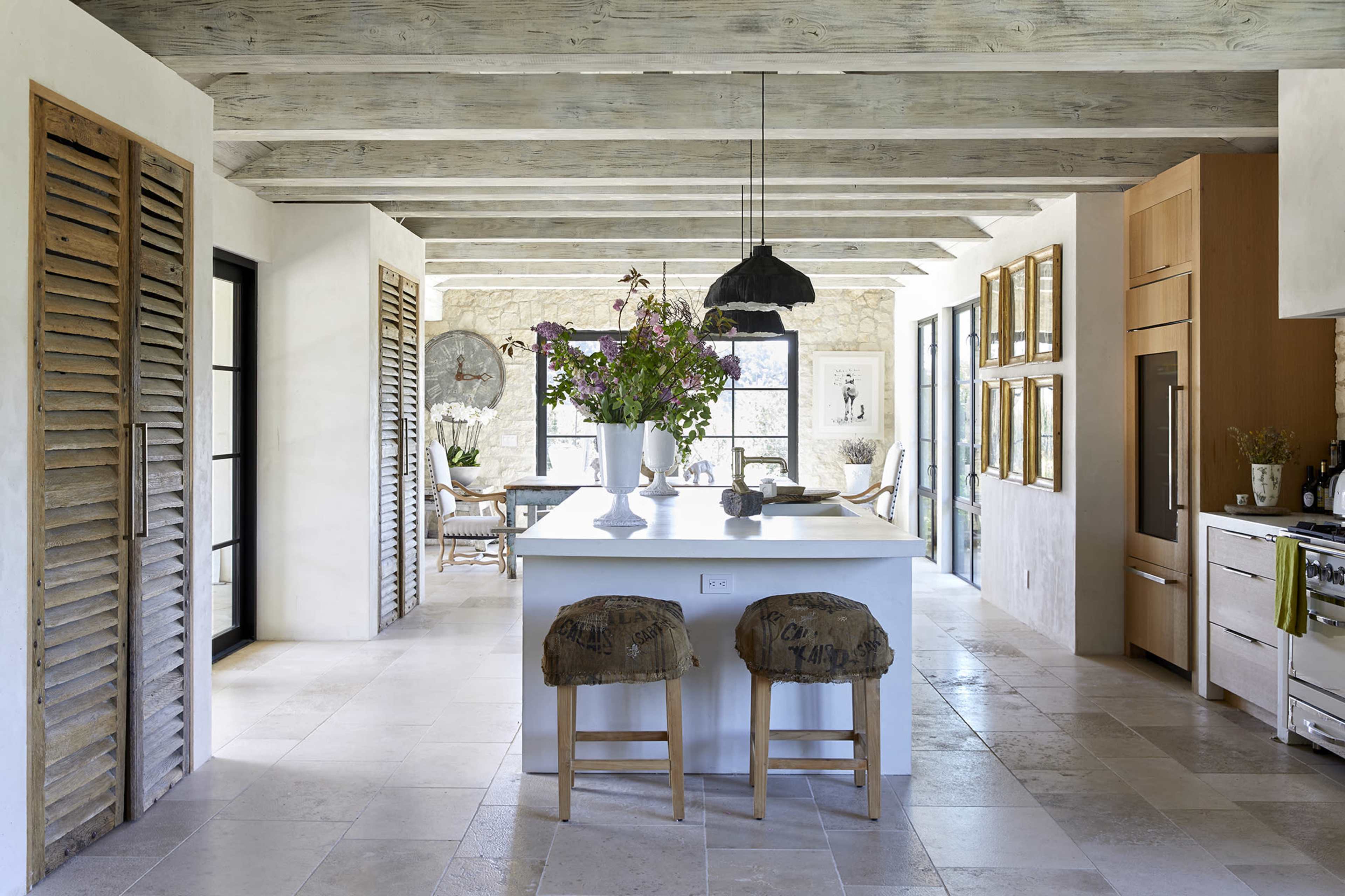 A spacious kitchen with a central island, adorned with a large vase of flowers, and flanked by stools, with wooden beams and stone walls in the background.