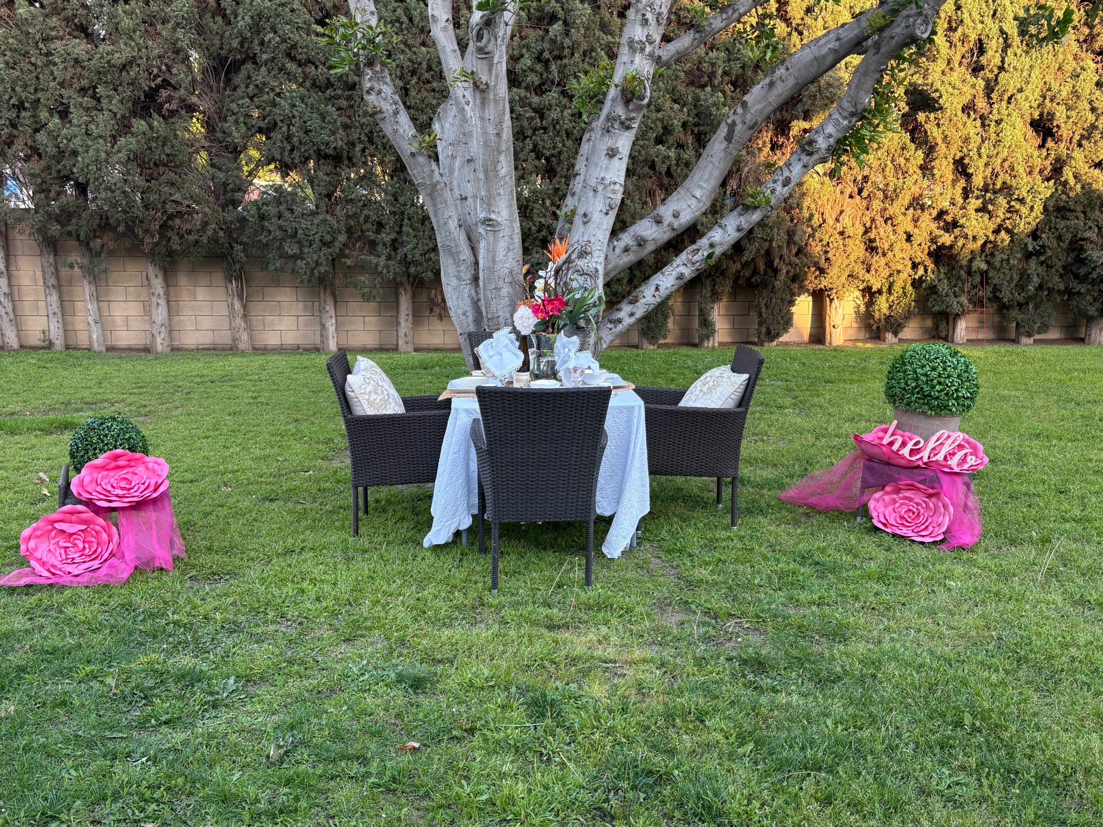 A dining table set for an outdoor gathering is surrounded by four wicker chairs and decorated with flowers and decorative greenery.