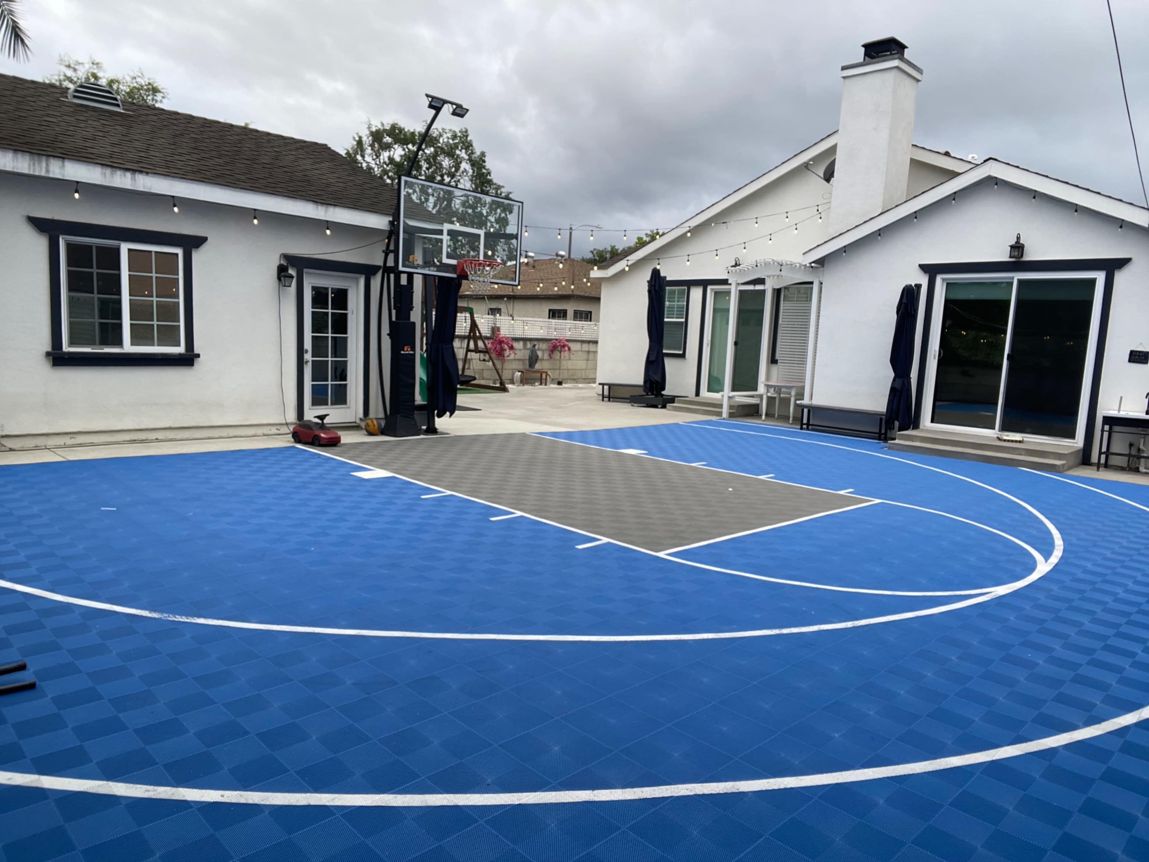The image shows a newly installed blue outdoor basketball court in a backyard, adjacent to a house with outdoor seating and a hoop on a backboard.