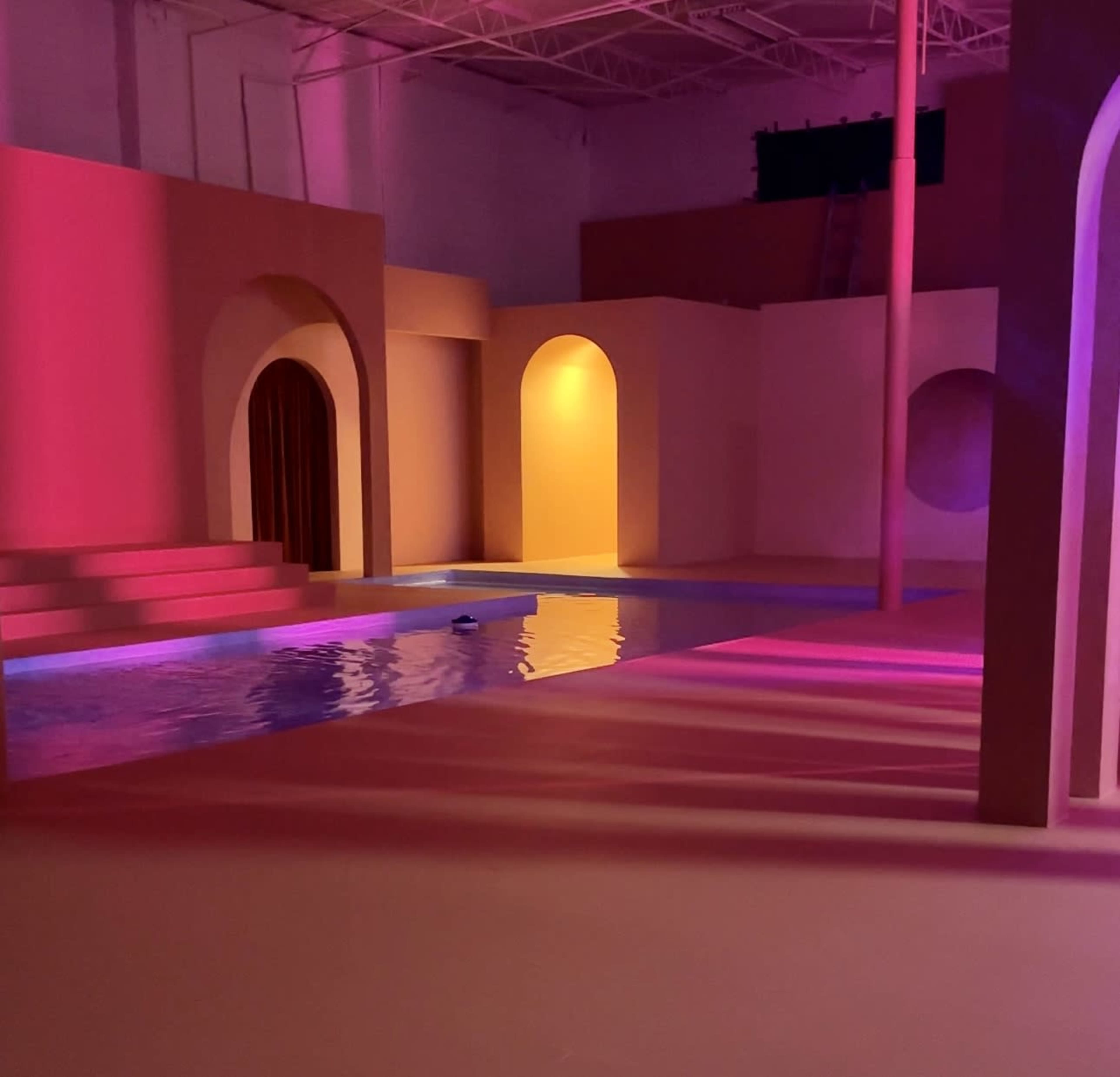 An indoor space featuring geometric arches, a small pool of water, and colored lighting.