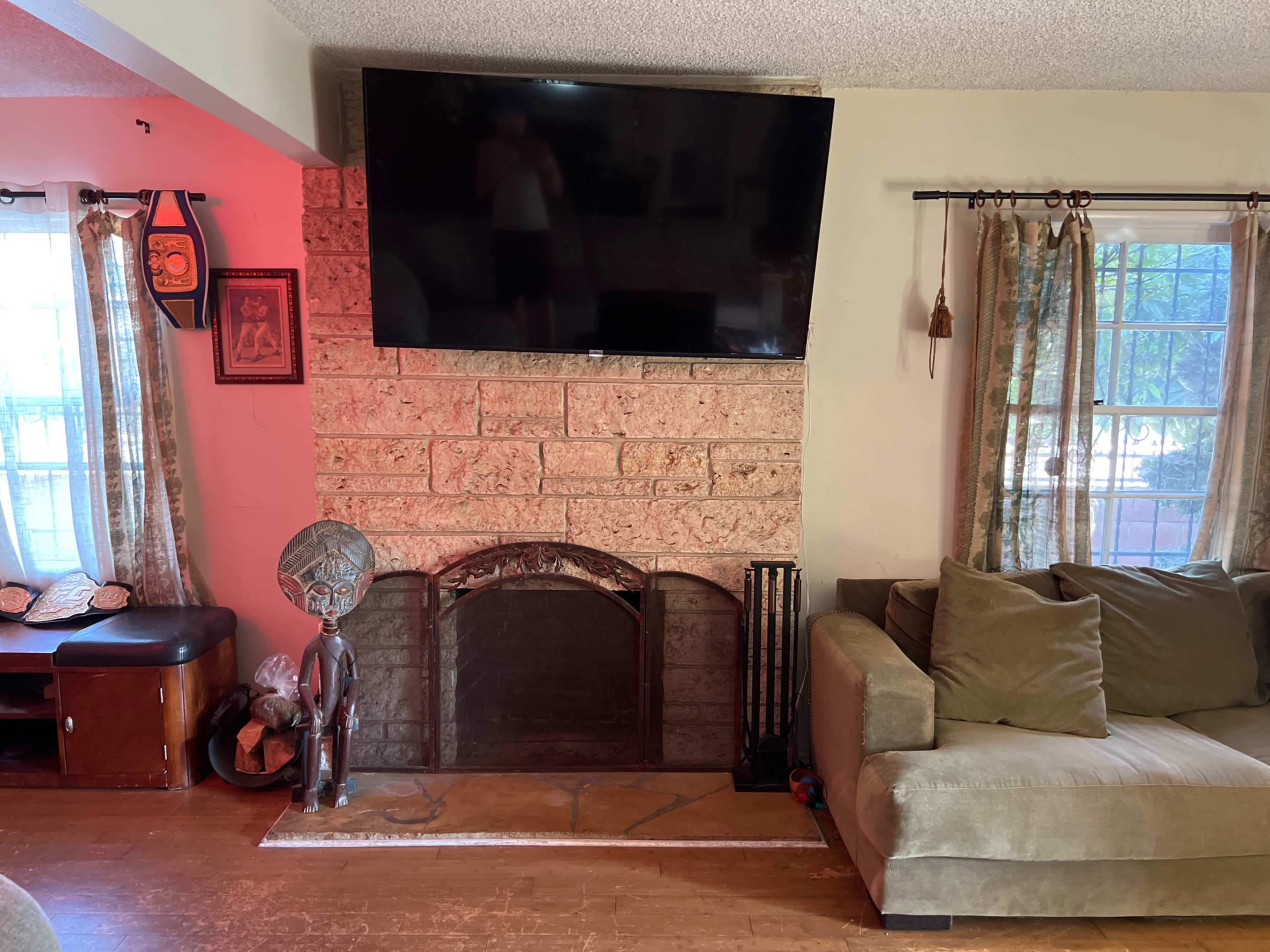 The image shows a living room with a stone fireplace featuring a large flat-screen TV mounted above it, a brown sofa on the right, and decorative items, including a statue and curtains, around the windows.